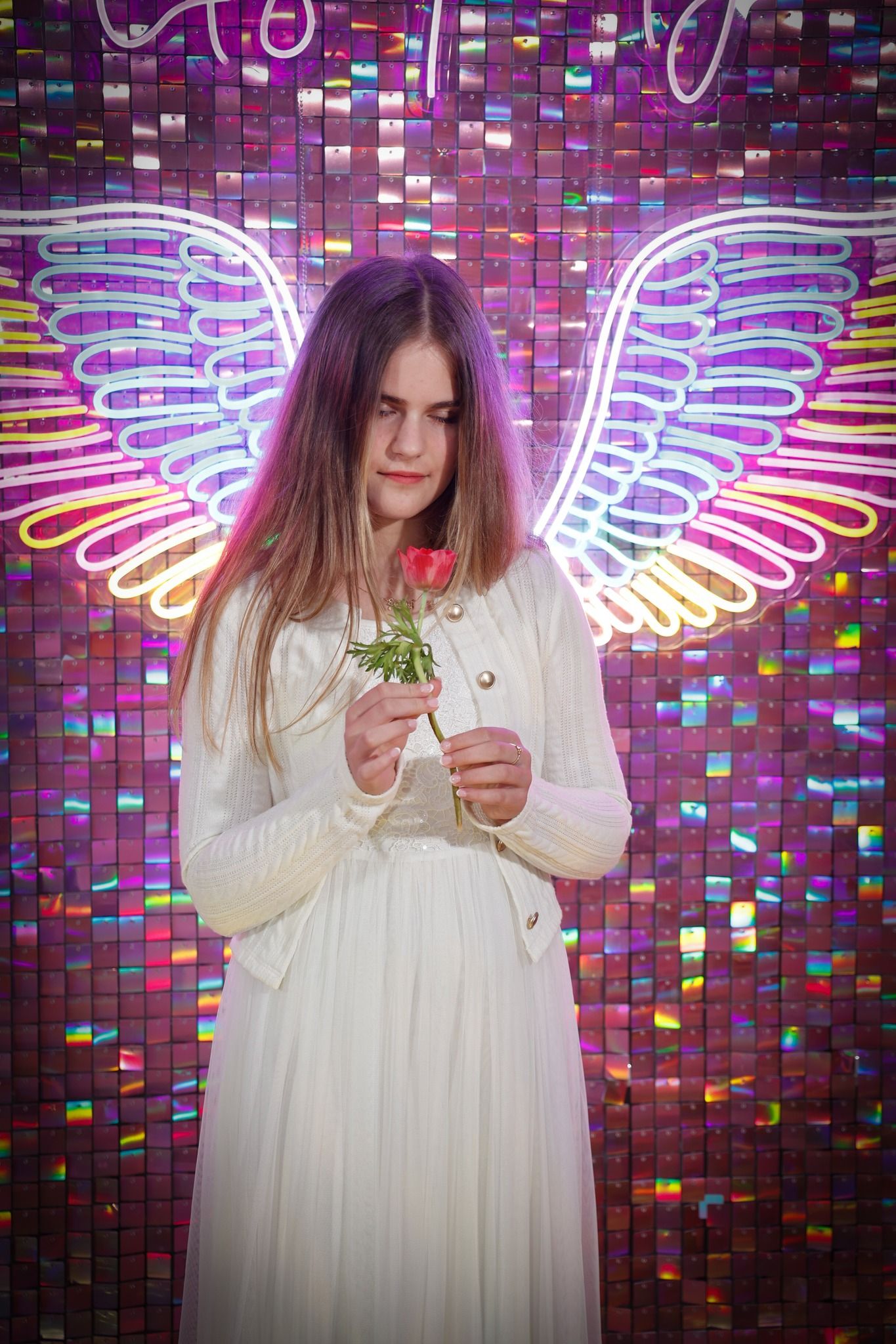 Person in white dress poses against neon angel wings wall art display.