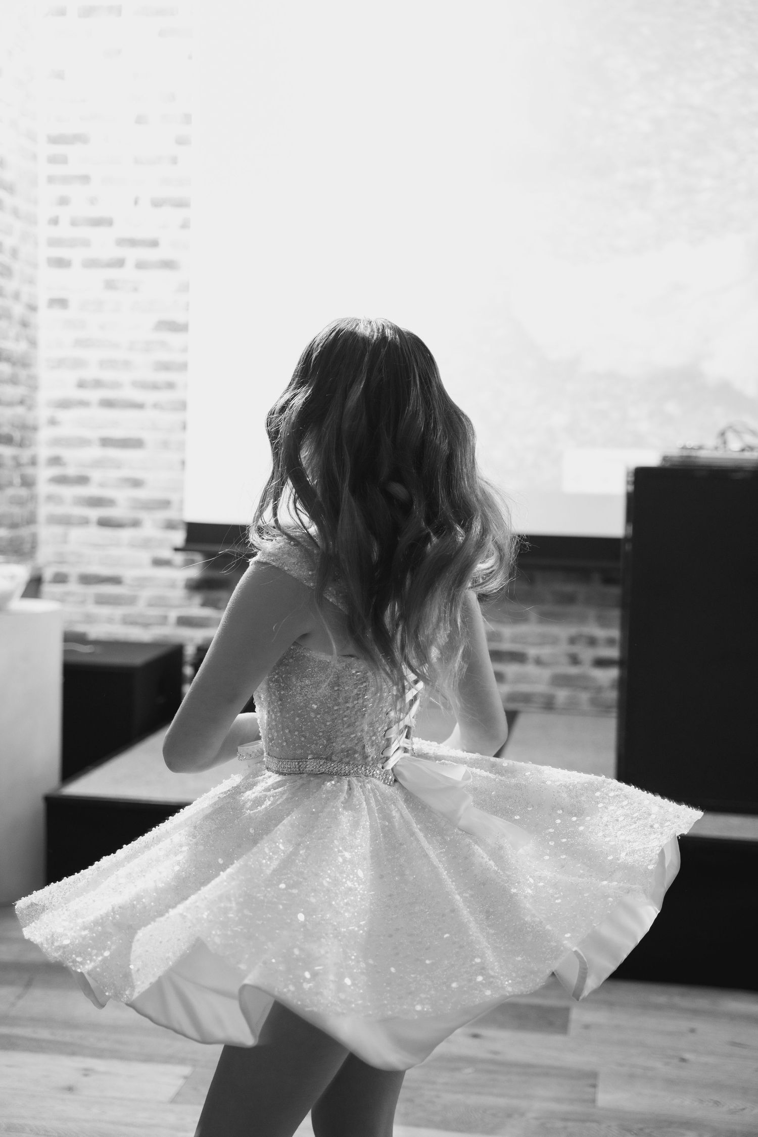 Young girl twirling in a sparkly white dress against a brick wall background.