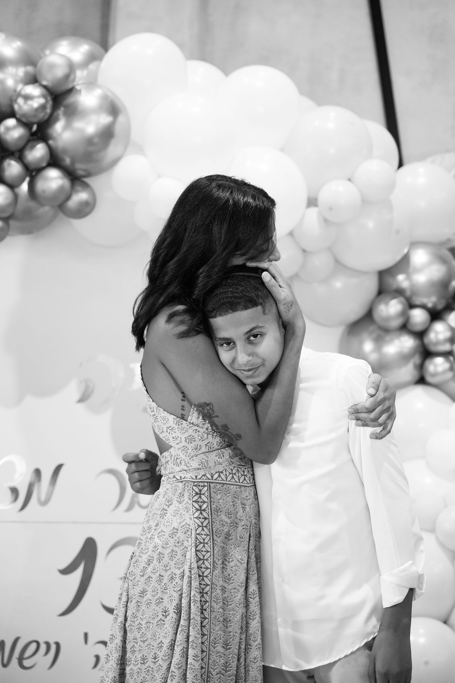 Young child in a white dress with embellished neckline poses against a backdrop of white and silver balloons.