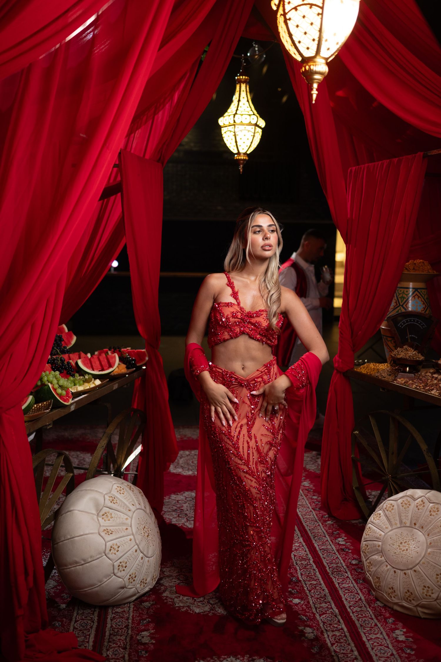 A woman in a red sequined two-piece outfit poses under draped red curtains with ornate lanterns and Moroccan decor.