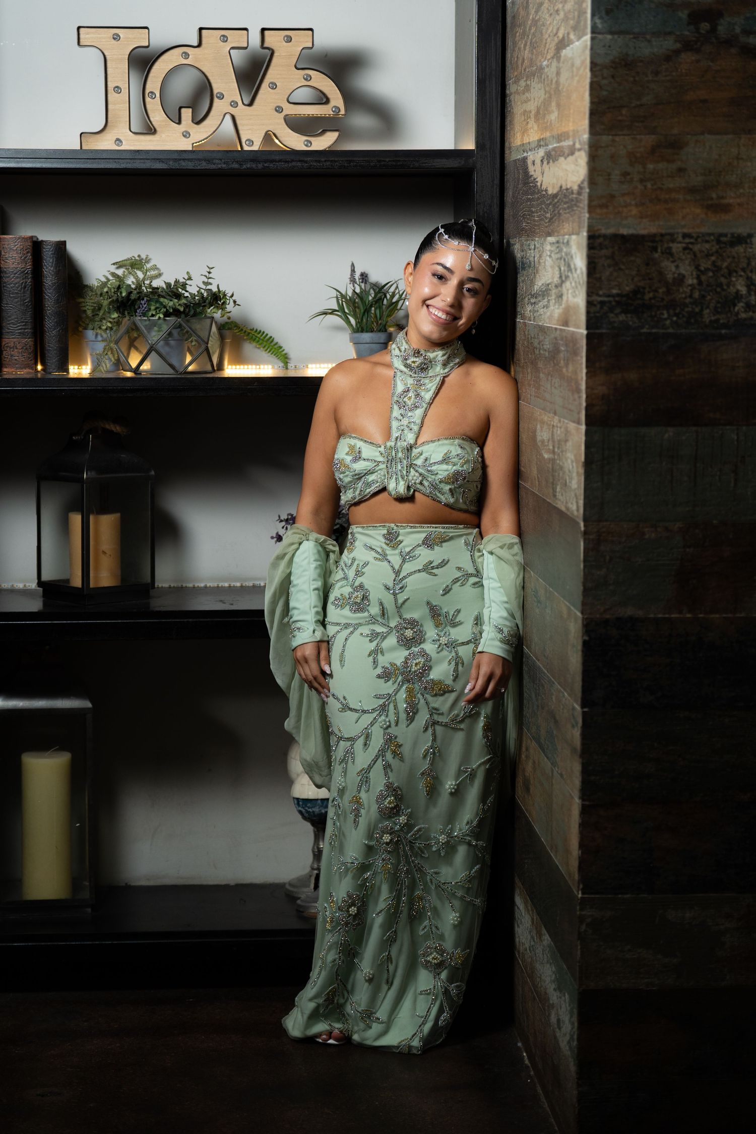 Woman in elegant sage green embroidered crop top and maxi skirt posing near shelves with a Love sign.