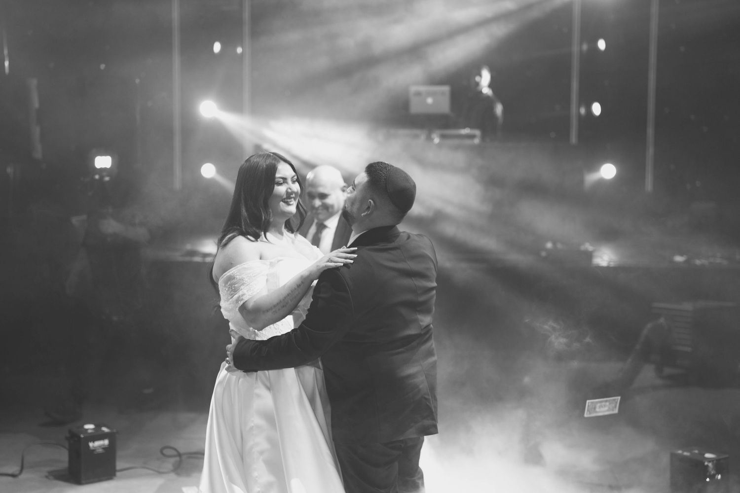 Black and white wedding photo of bride and groom sharing first dance under dramatic stage lighting.
