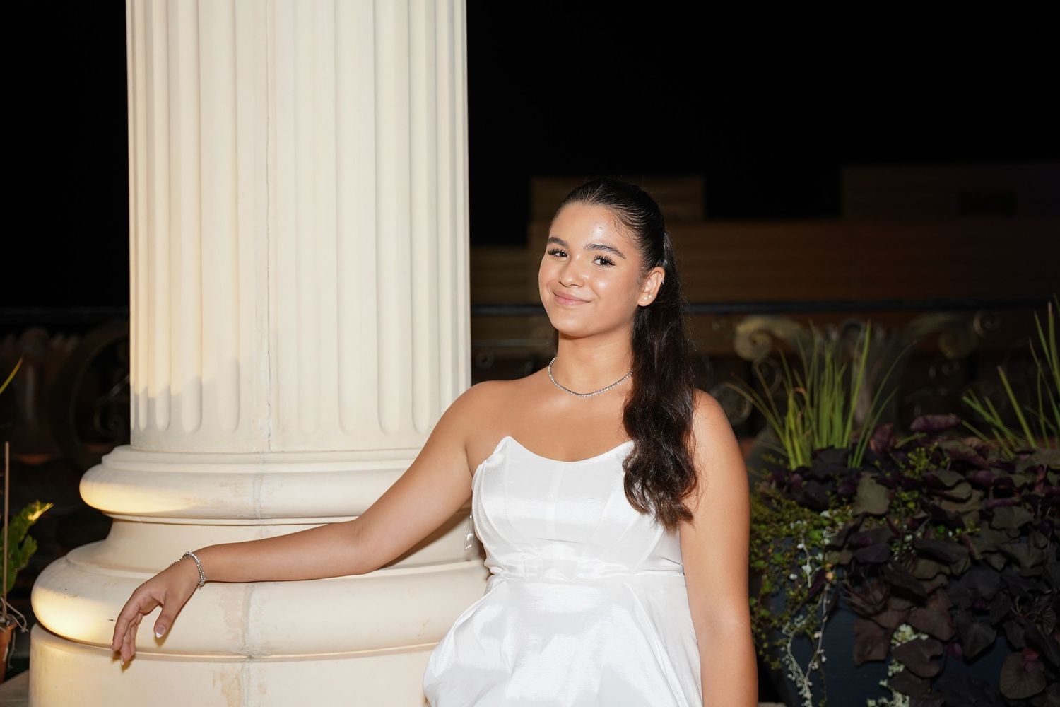 Woman in white dress posing elegantly against a classic white column outdoors.