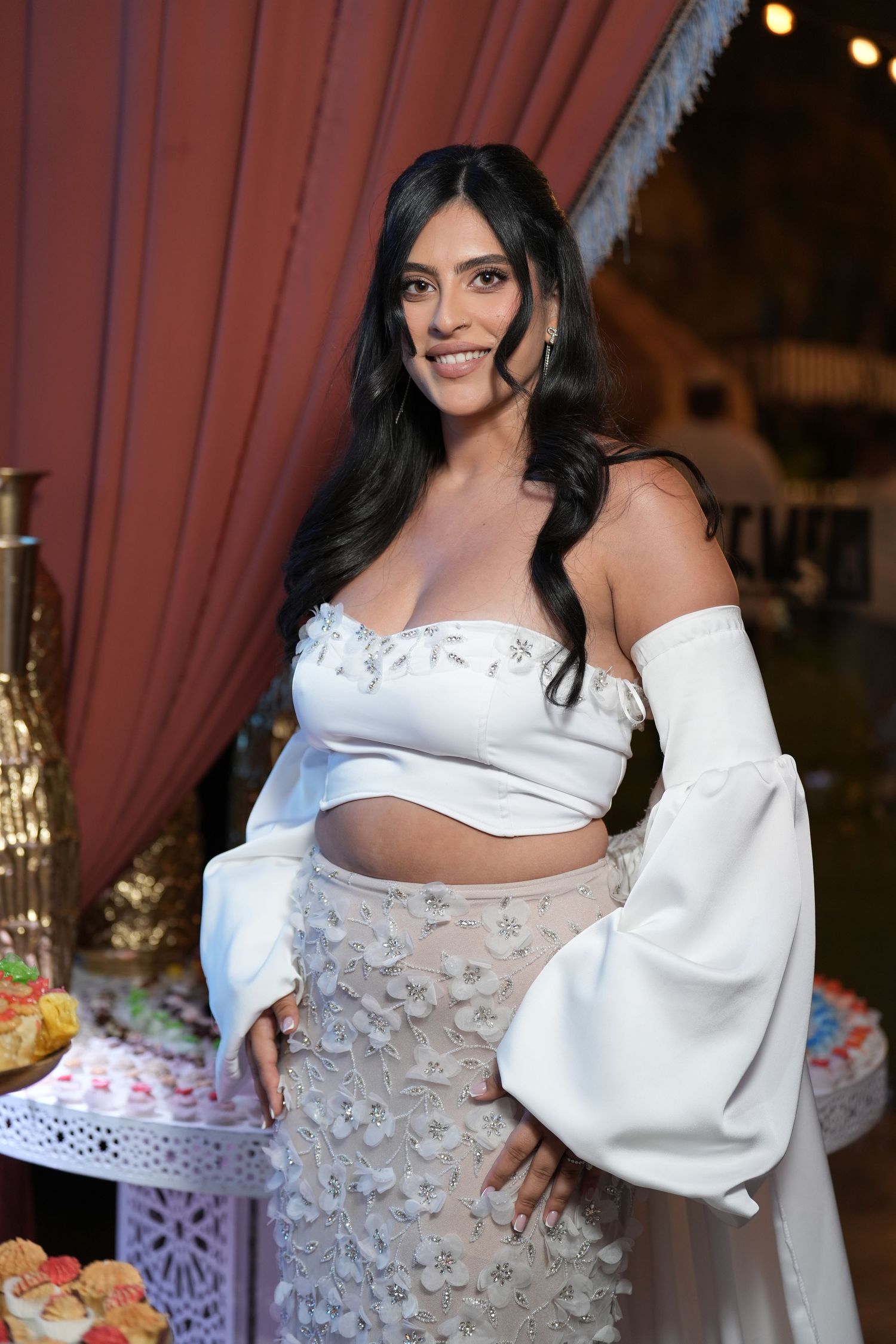 Woman in white off-shoulder crop top and floral skirt smiling at elegant event with dessert table backdrop.