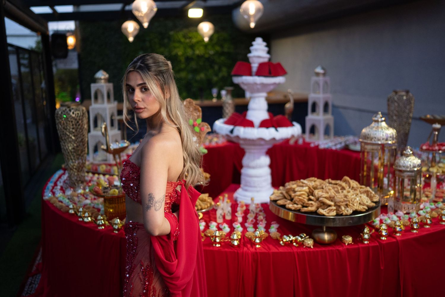 Woman in red gown poses at elegant dessert table with red and gold decorations and a white fountain centerpiece.