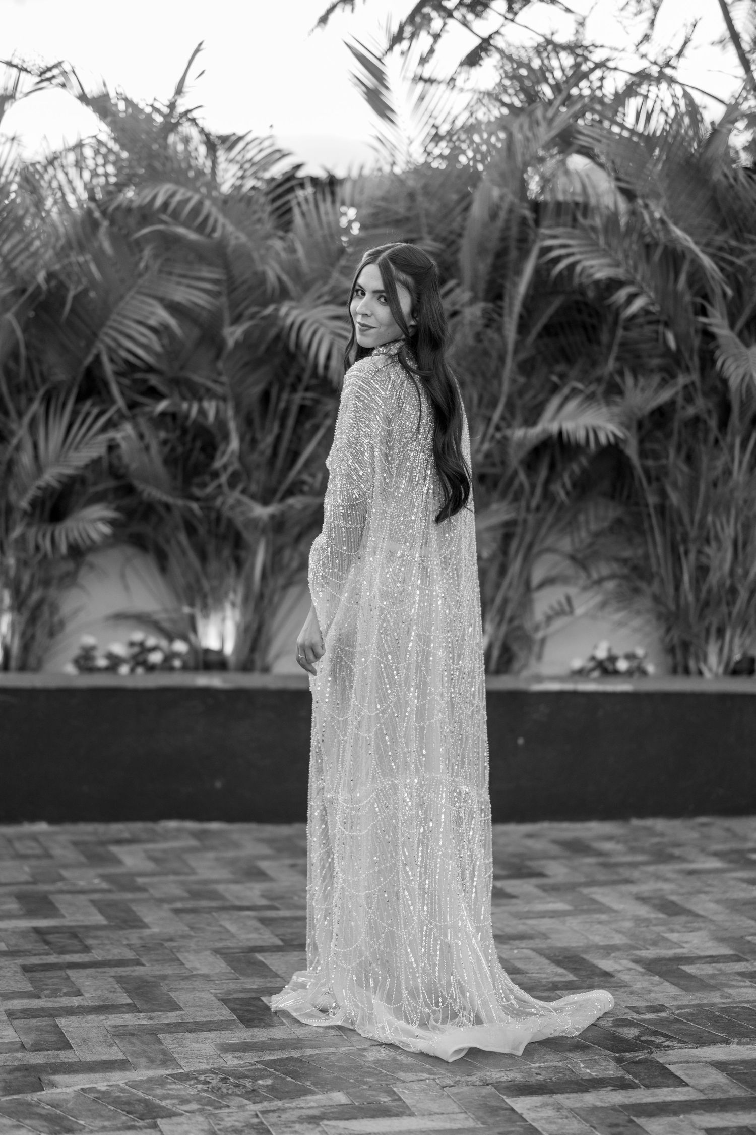 Black and white photo of a bride in a beaded gown with cape, posing before lush tropical palms.
