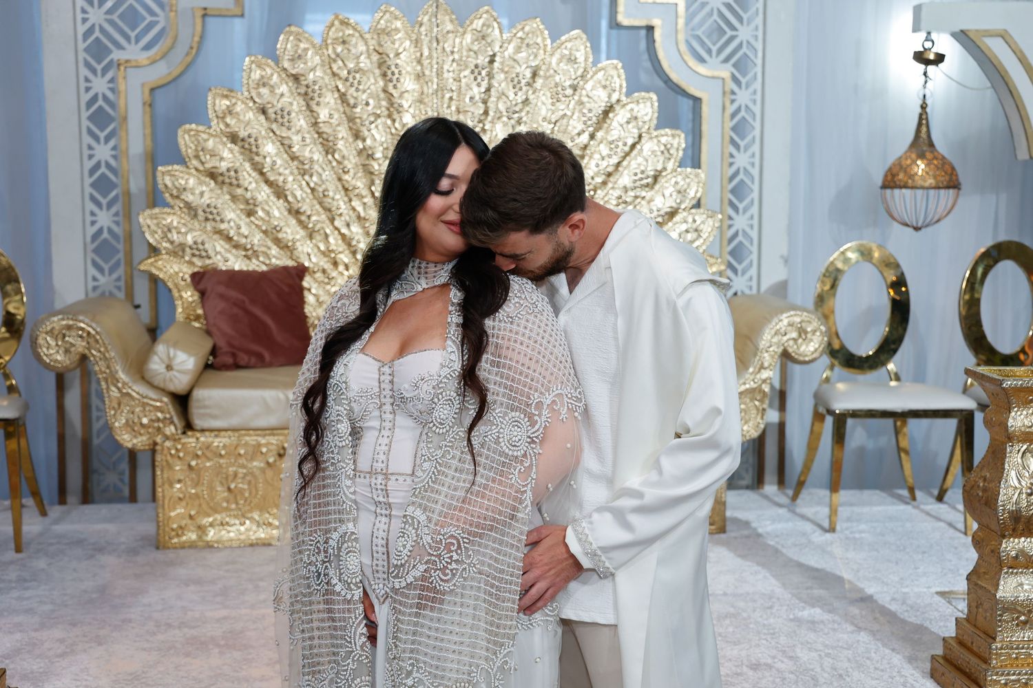 A couple in white attire posing against an ornate gold fan backdrop.