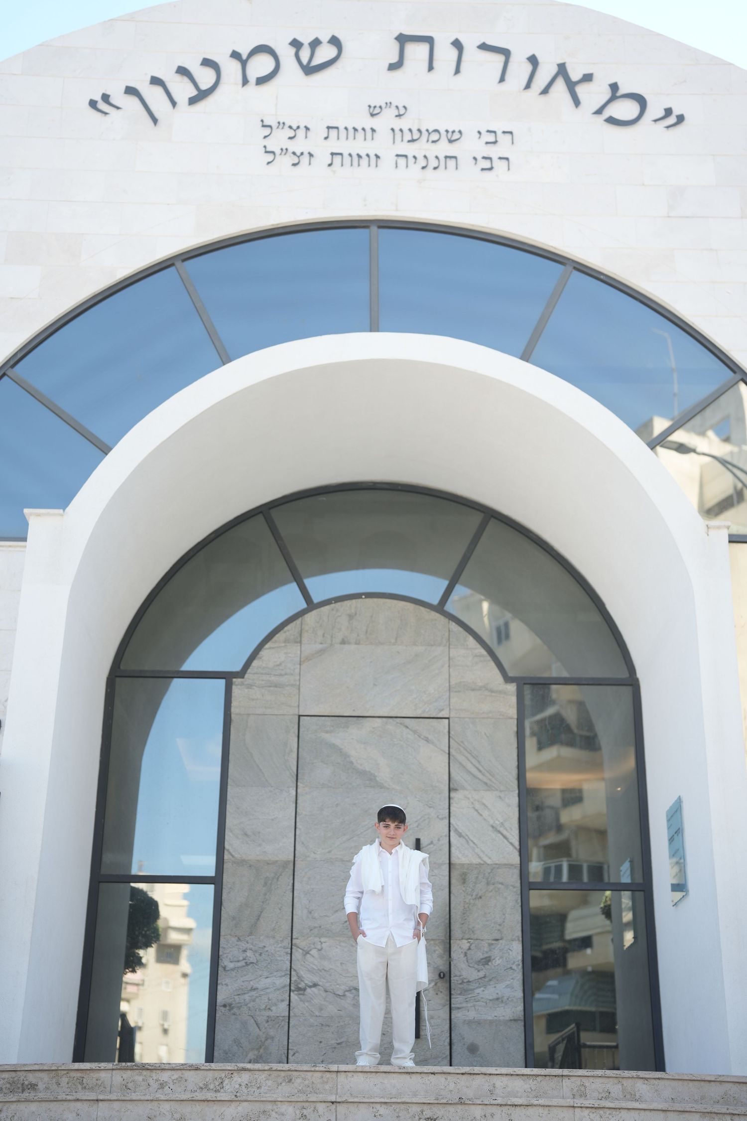 Boy in white outfit standing at entrance of Mearat Shimon synagogue with Hebrew inscription above arch.