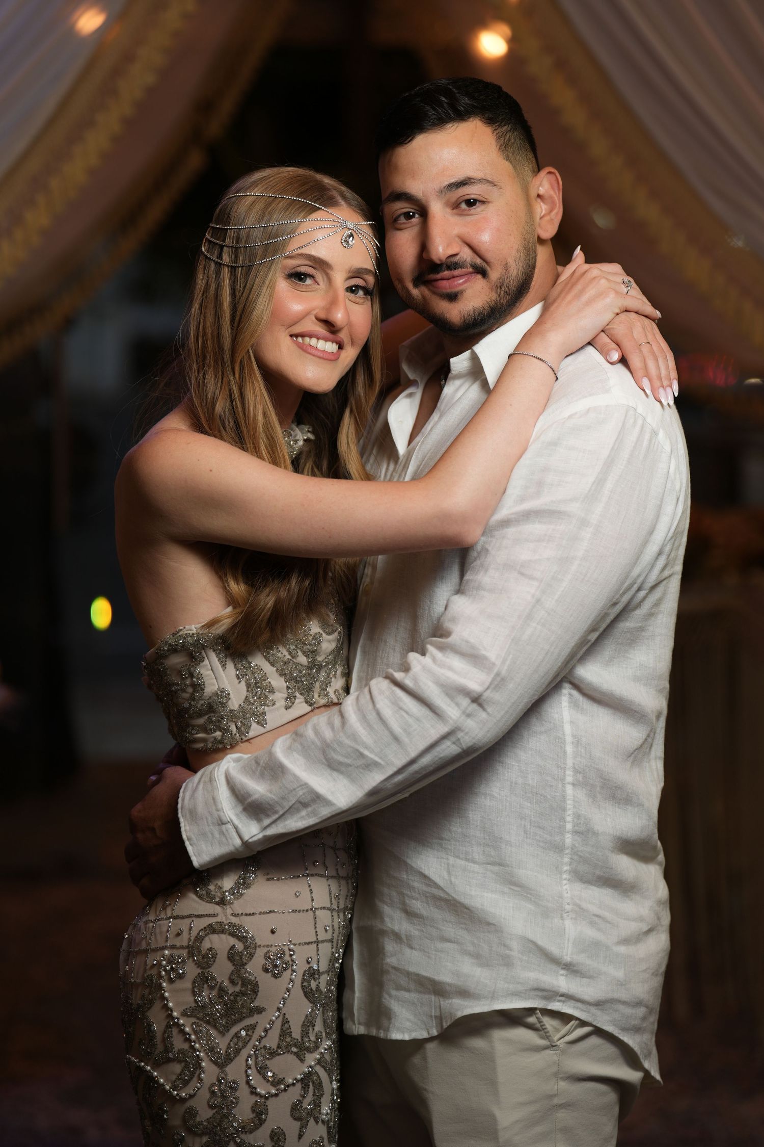 Smiling couple in elegant attire embracing at a warmly lit event, woman wearing beaded dress and headpiece.