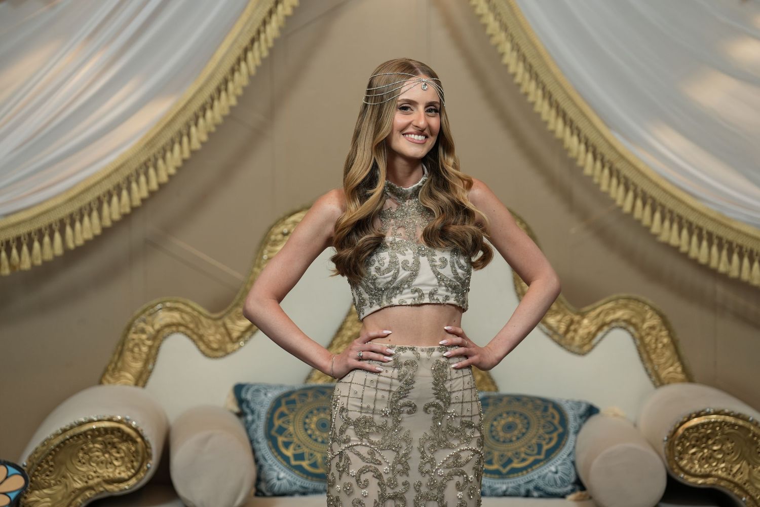 Young woman in elegant silver crop top and skirt ensemble posing confidently in ornate gold and white venue.