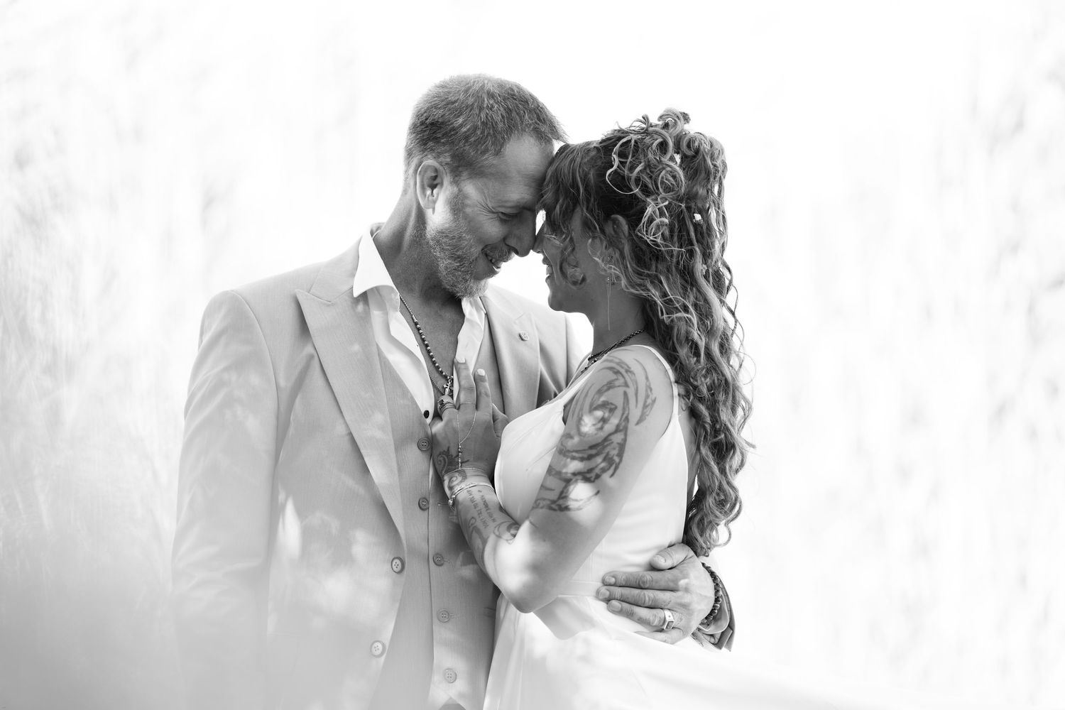 Black and white wedding photo of a couple in an intimate embrace, bride in white gown with tattoo, groom in light suit.