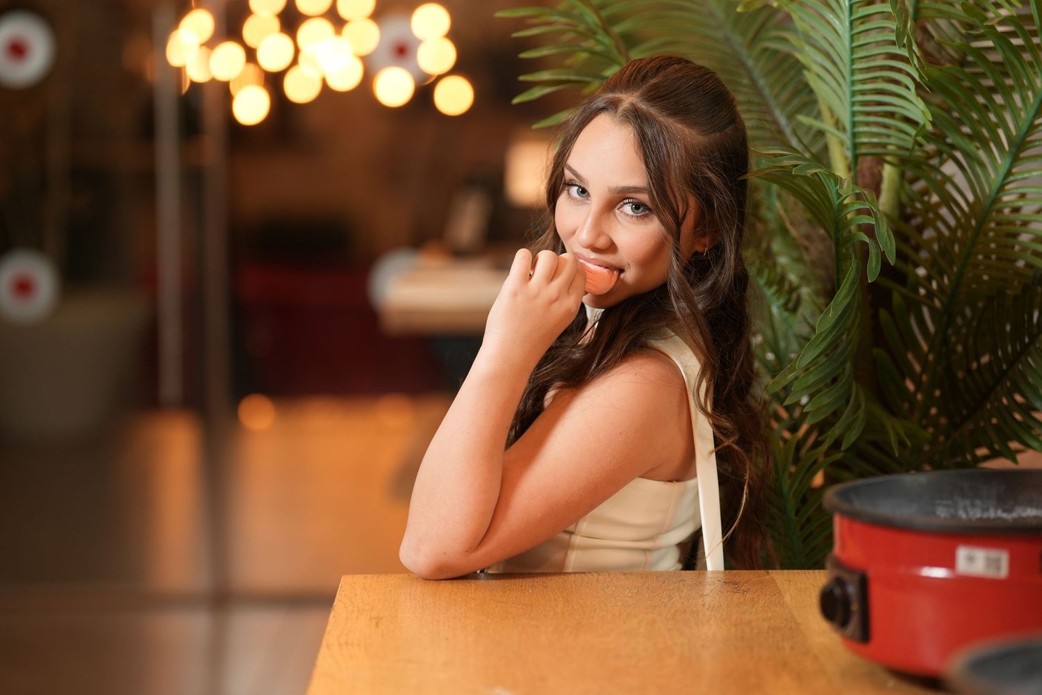 Young brunette woman sitting at a wooden table in a restaurant, smiling over her shoulder near a green plant.