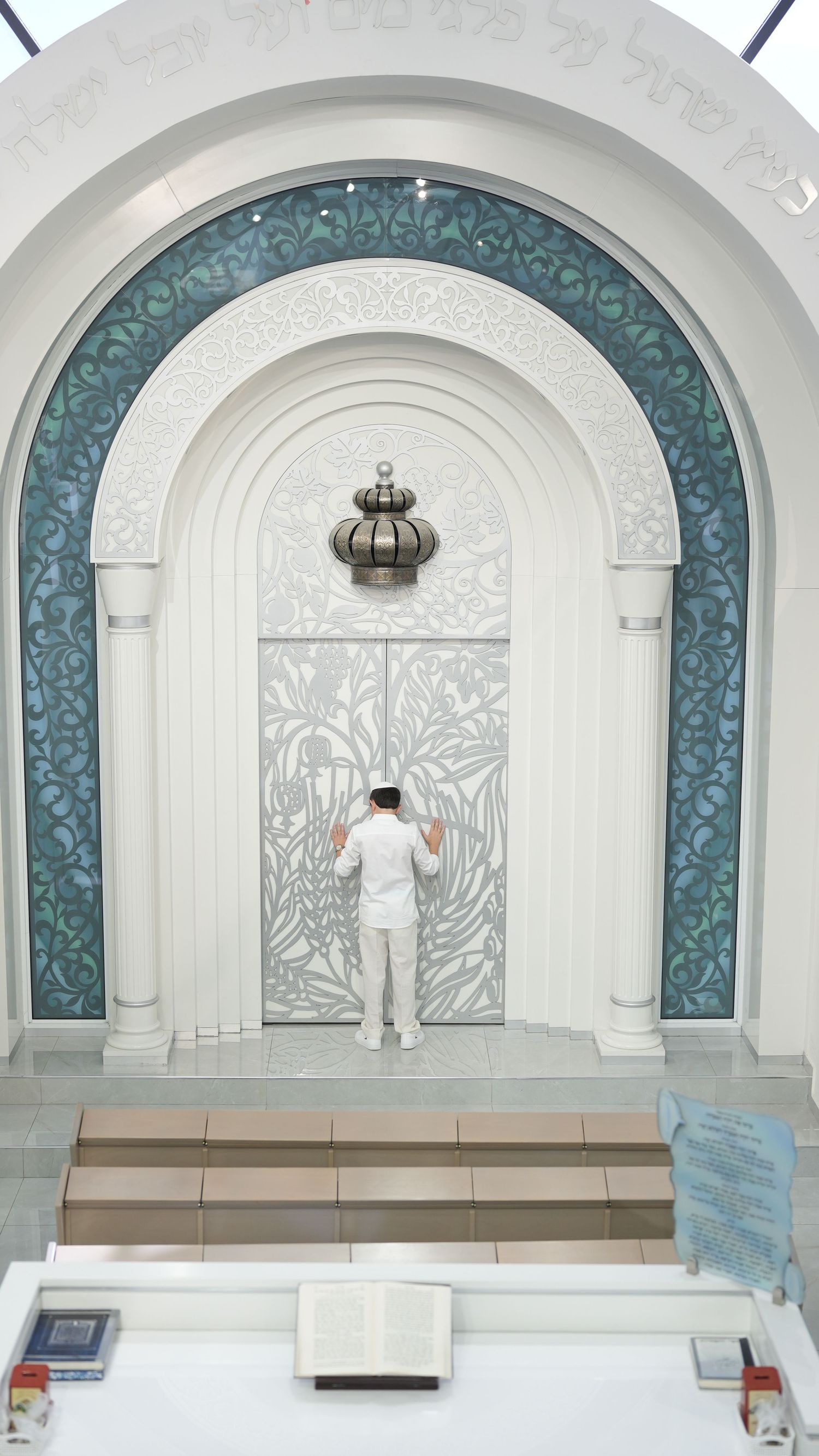 A man in white attire prays before an ornate white arched doorway with Hebrew inscriptions in a synagogue.