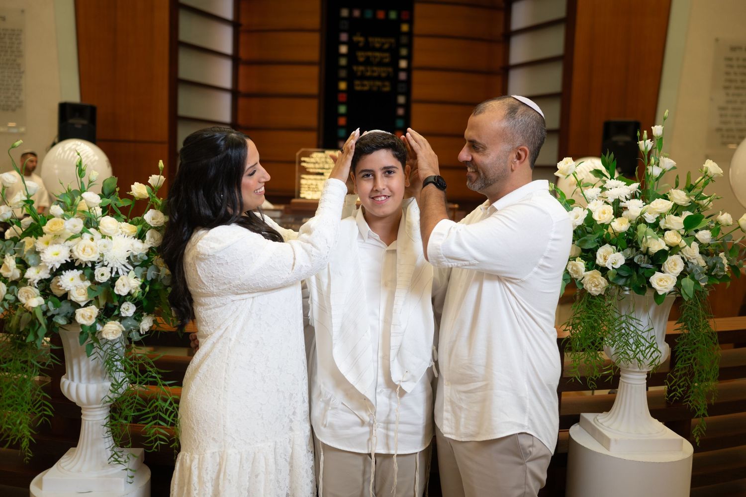 People in white attire standing together near elegant floral decorations at an indoor venue.