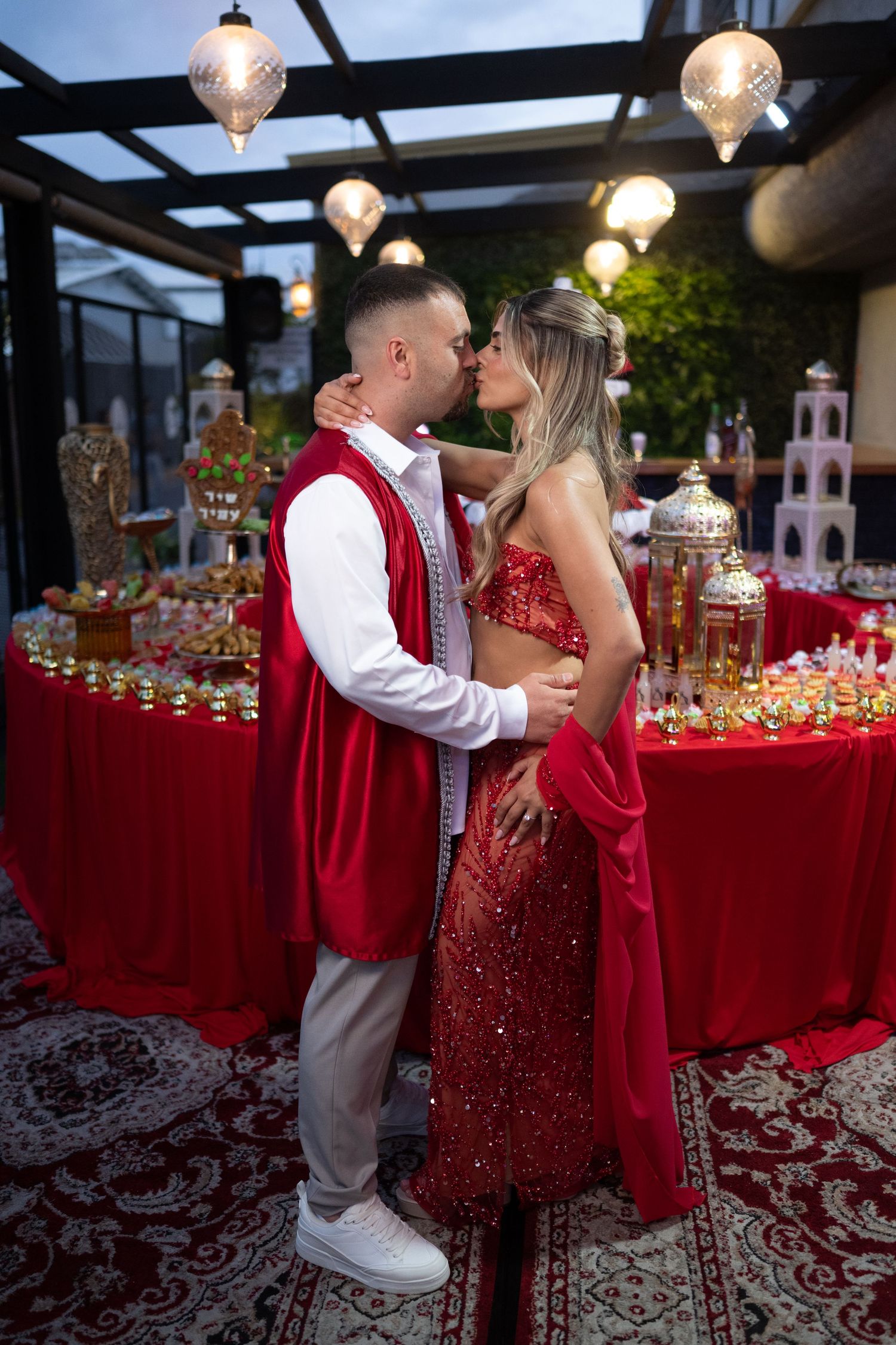 Couple kissing in red festive outfits at elegant decorated party venue with lanterns and red table display.