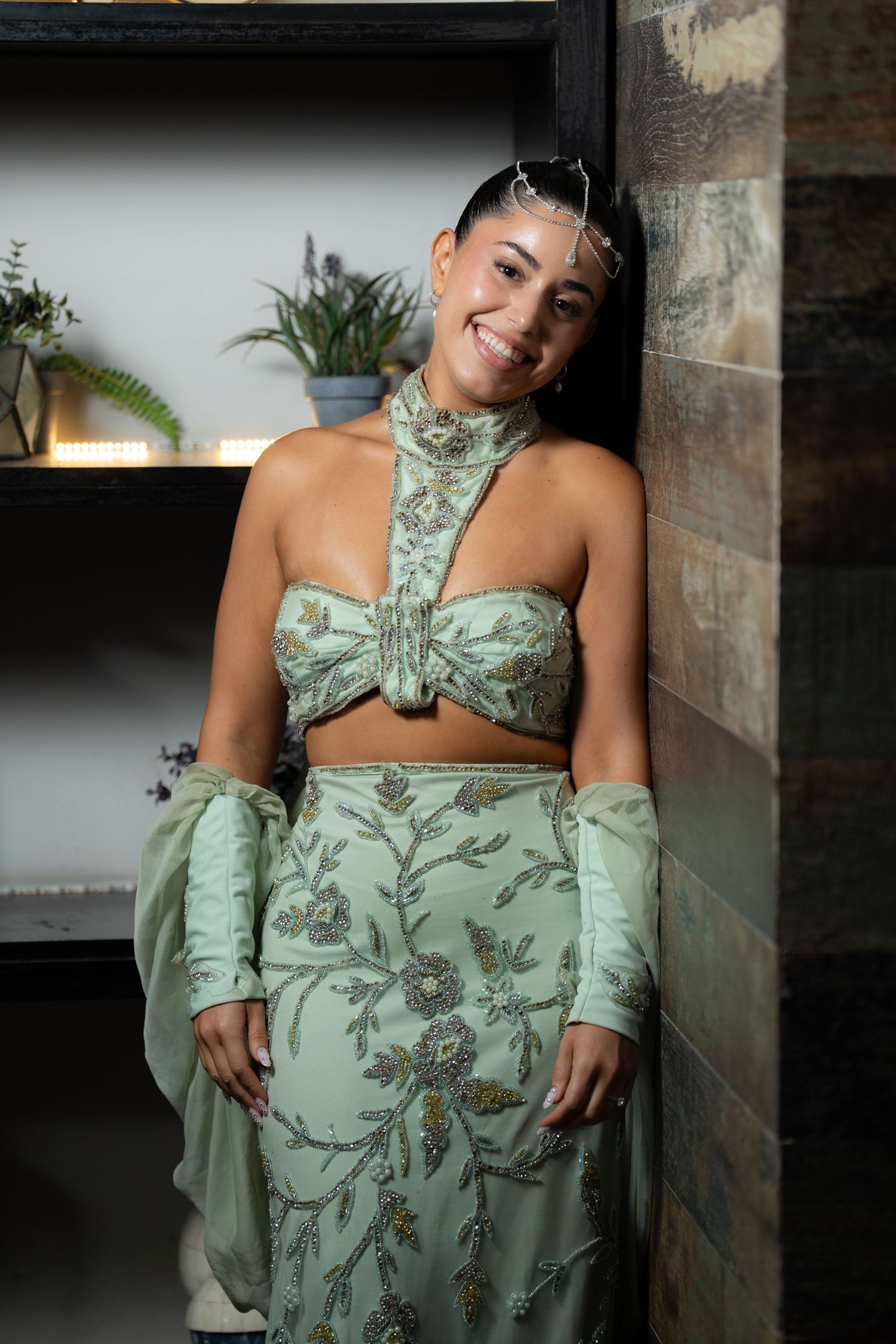 A smiling woman poses in an elegant sage green embroidered crop top and skirt set with floral beading, leaning against a wall.