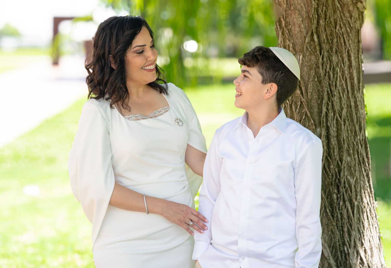 A couple in white outfits shares tender moments in a sunlit park setting with green trees in the background.
