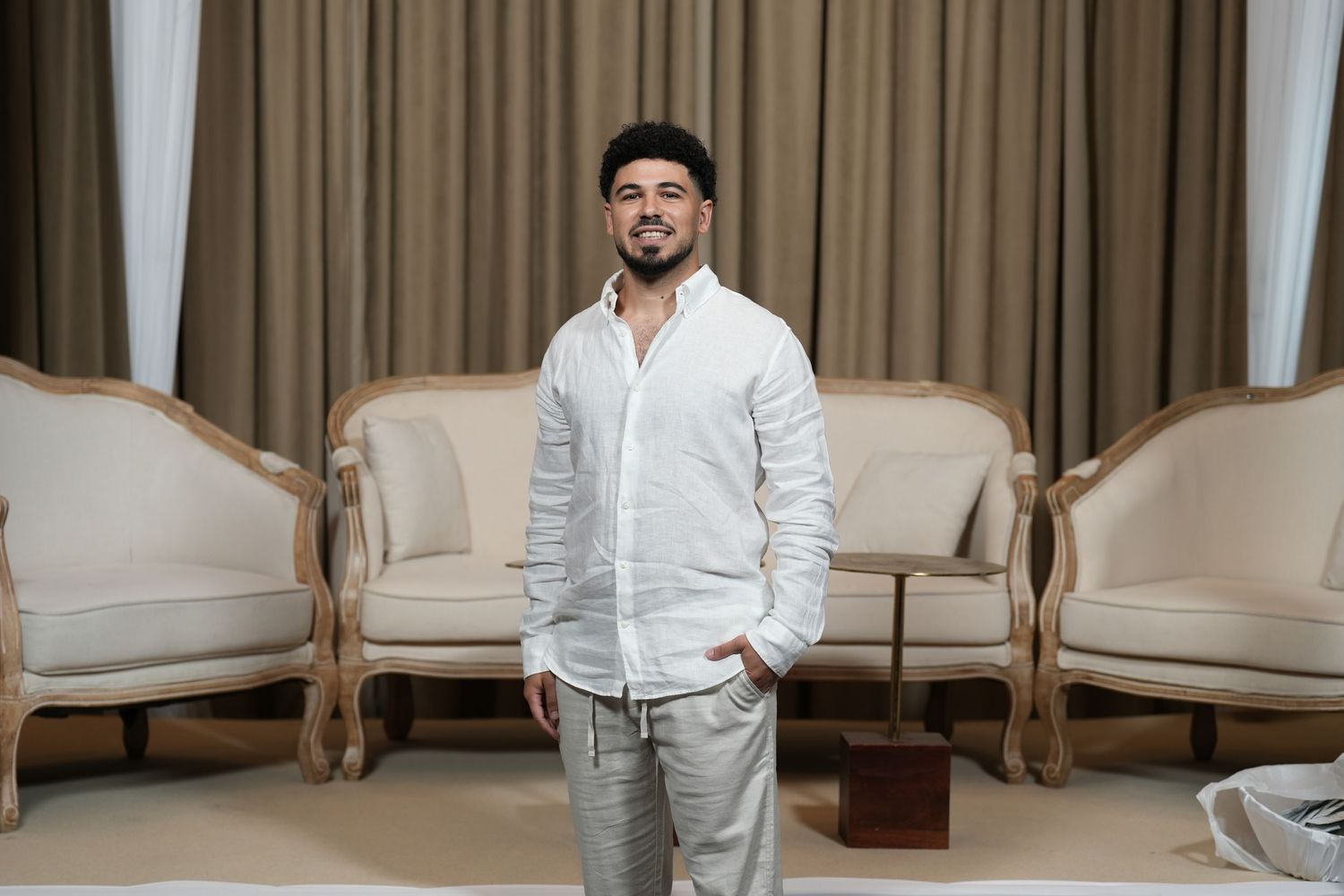 A young man in a white linen shirt and gray pants stands confidently between two elegant cream chairs.