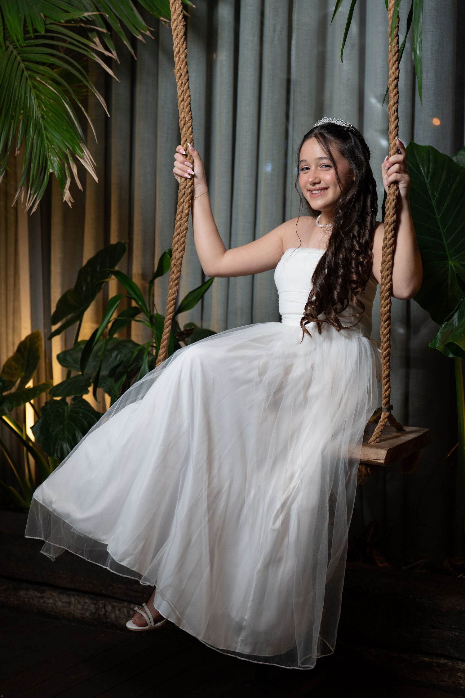 A person in a flowing white dress swings playfully on a rope swing against a dark backdrop with tropical plants.