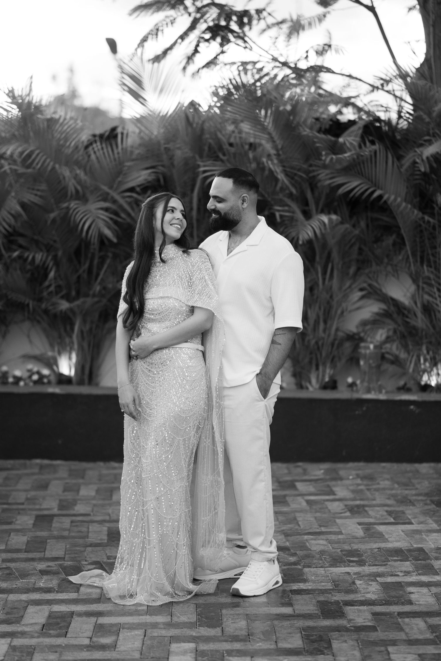 Black and white photo of a smiling pregnant couple posing outdoors with tropical palm trees in the background.