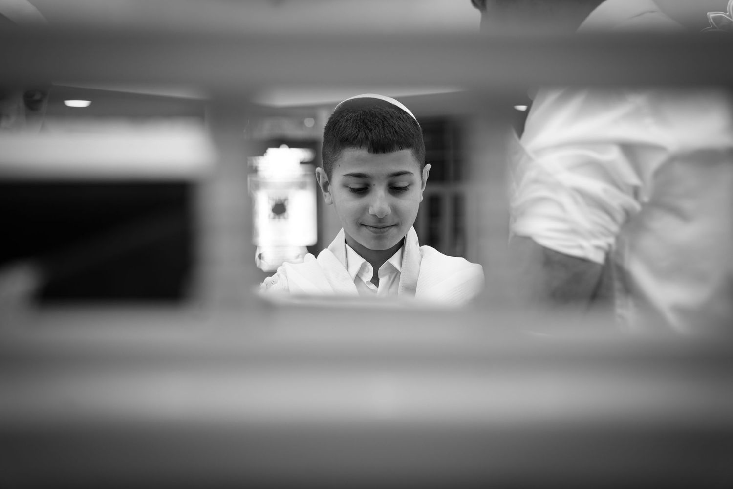 Black and white photo of a young boy wearing a kippah, looking down thoughtfully in a formal setting.