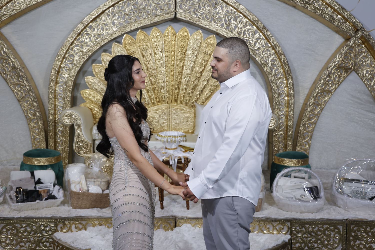A couple holds hands during a wedding ceremony in front of an ornate gold backdrop with decorative table settings.