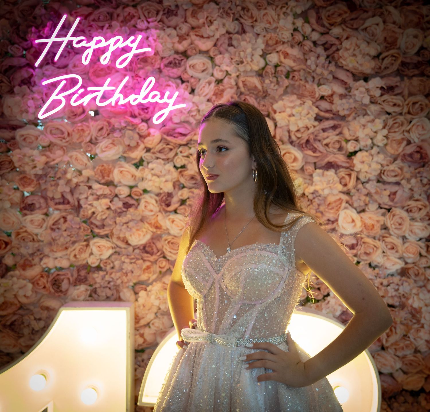 White lace dress against floral backdrop with pink neon 'Happy Birthday' sign and dramatic lighting.