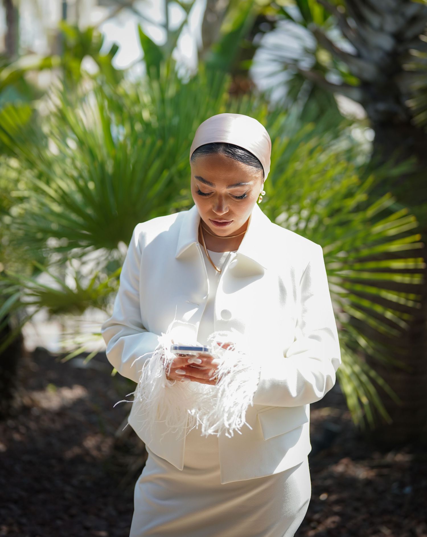 Person in white attire checking phone while sitting among tropical palm plants.