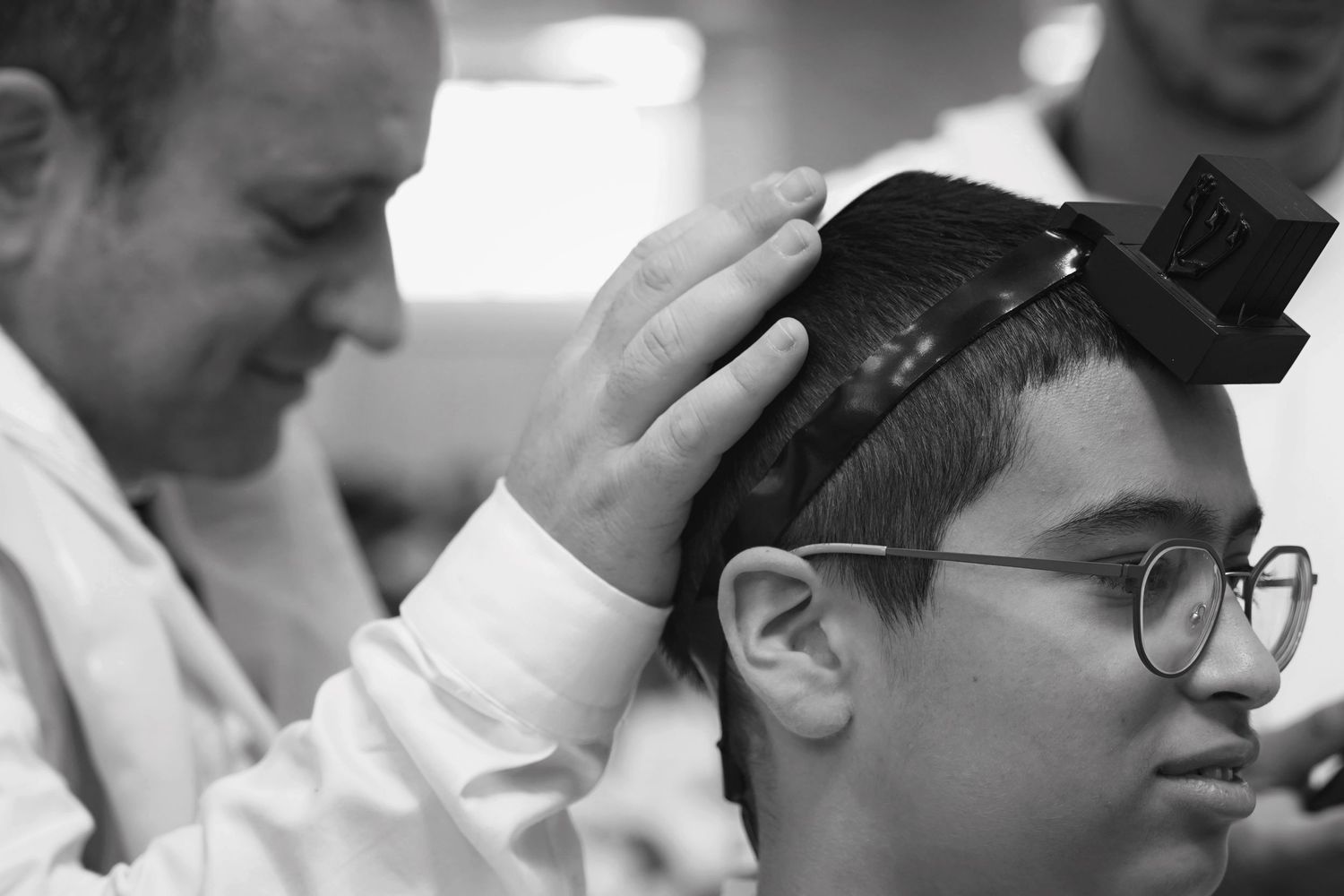 A close-up black and white image of someone adjusting a small hat or headpiece on another person wearing glasses.
