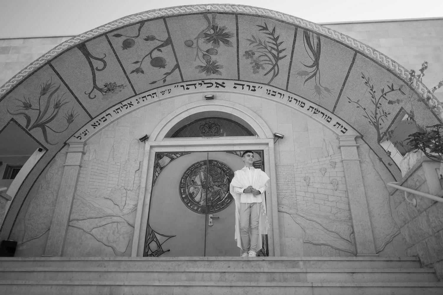 A black and white photo of someone posing with fabric wings under a decorated archway.