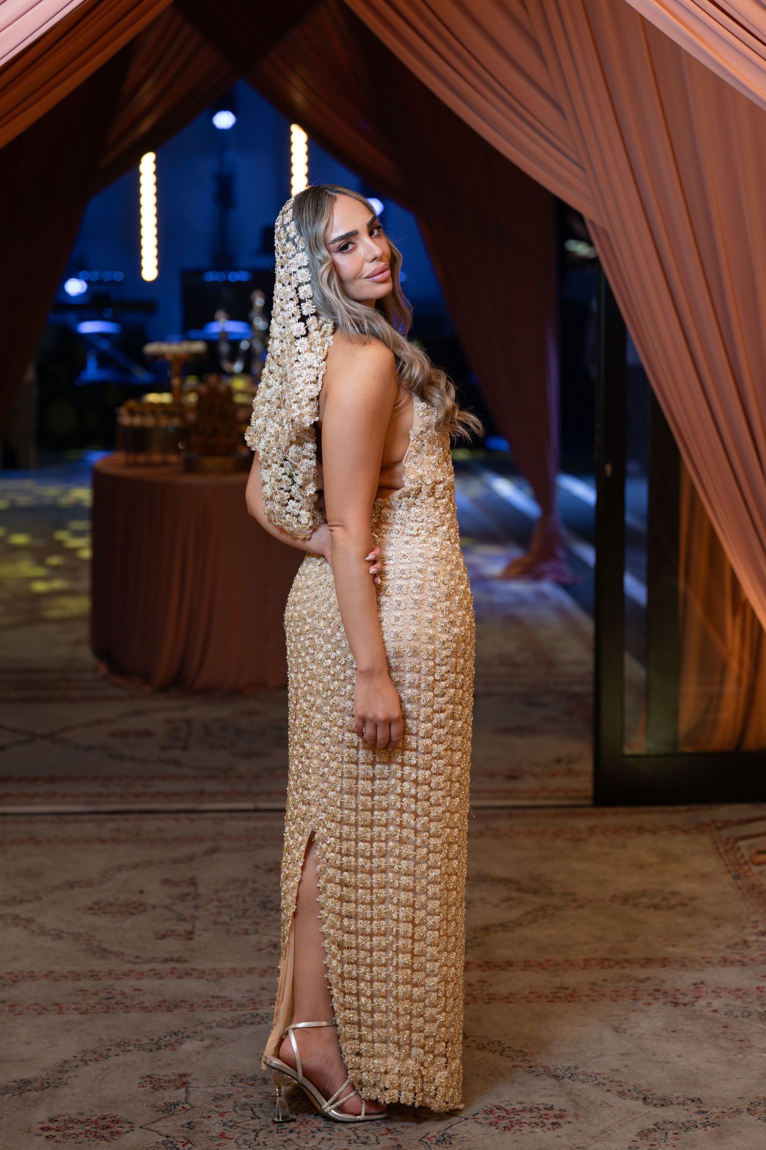 Woman in gold sequin gown with floral headpiece poses elegantly at a formal event.