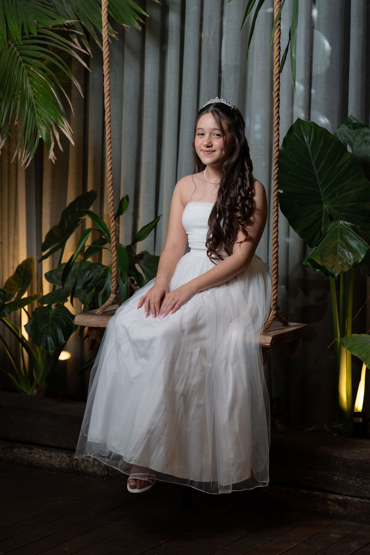 A person in a white tulle wedding dress sits gracefully on a swing with tropical plants in the background.
