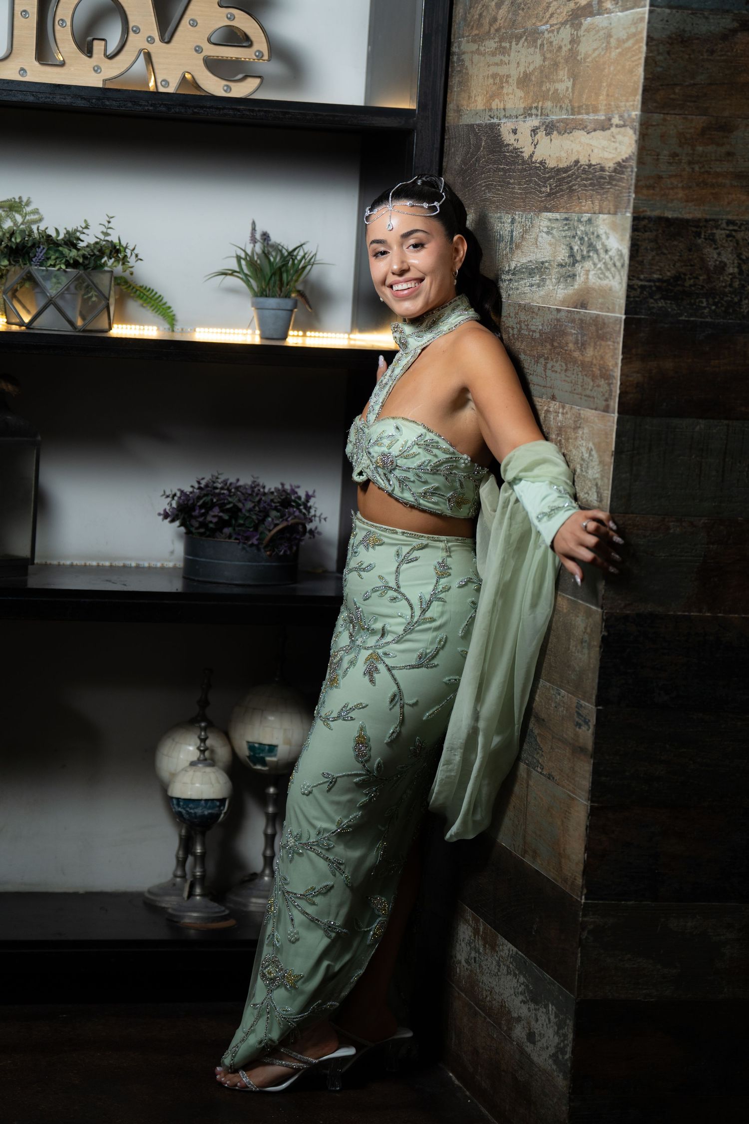 Woman in elegant sage green floral crop top and skirt set, posing gracefully beside a rustic wooden shelf with plants.