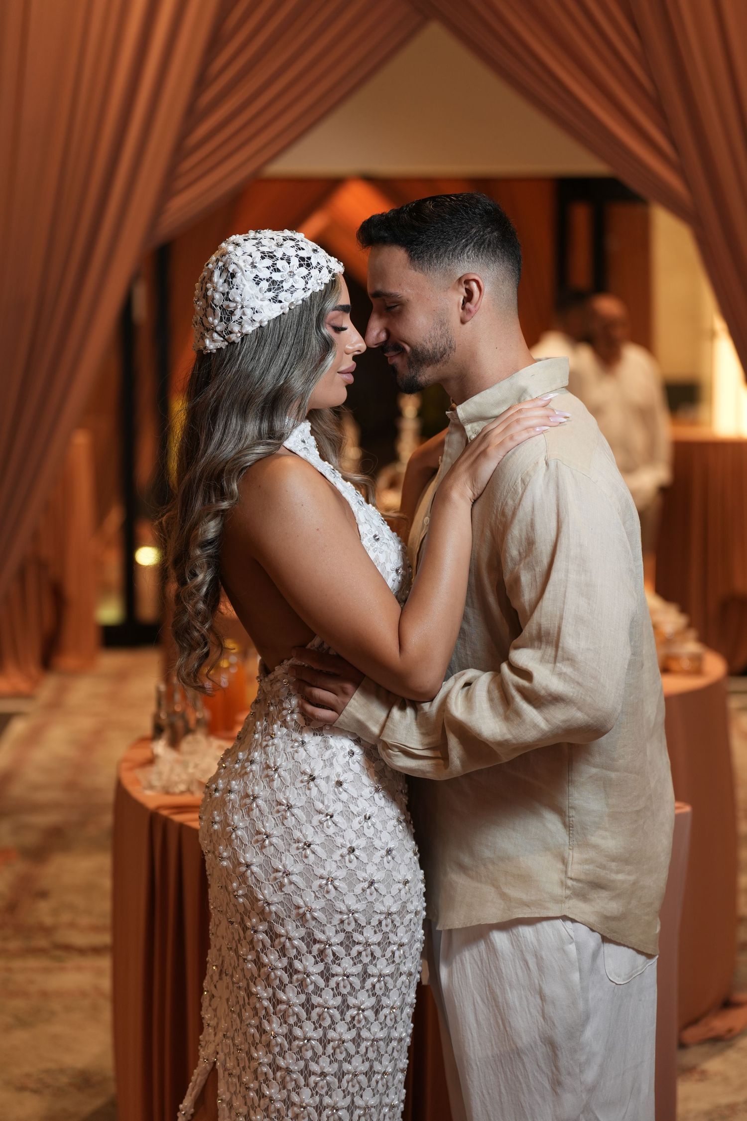 Couple in elegant wedding attire sharing a romantic moment, woman in white lace dress and headpiece, man in beige outfit.