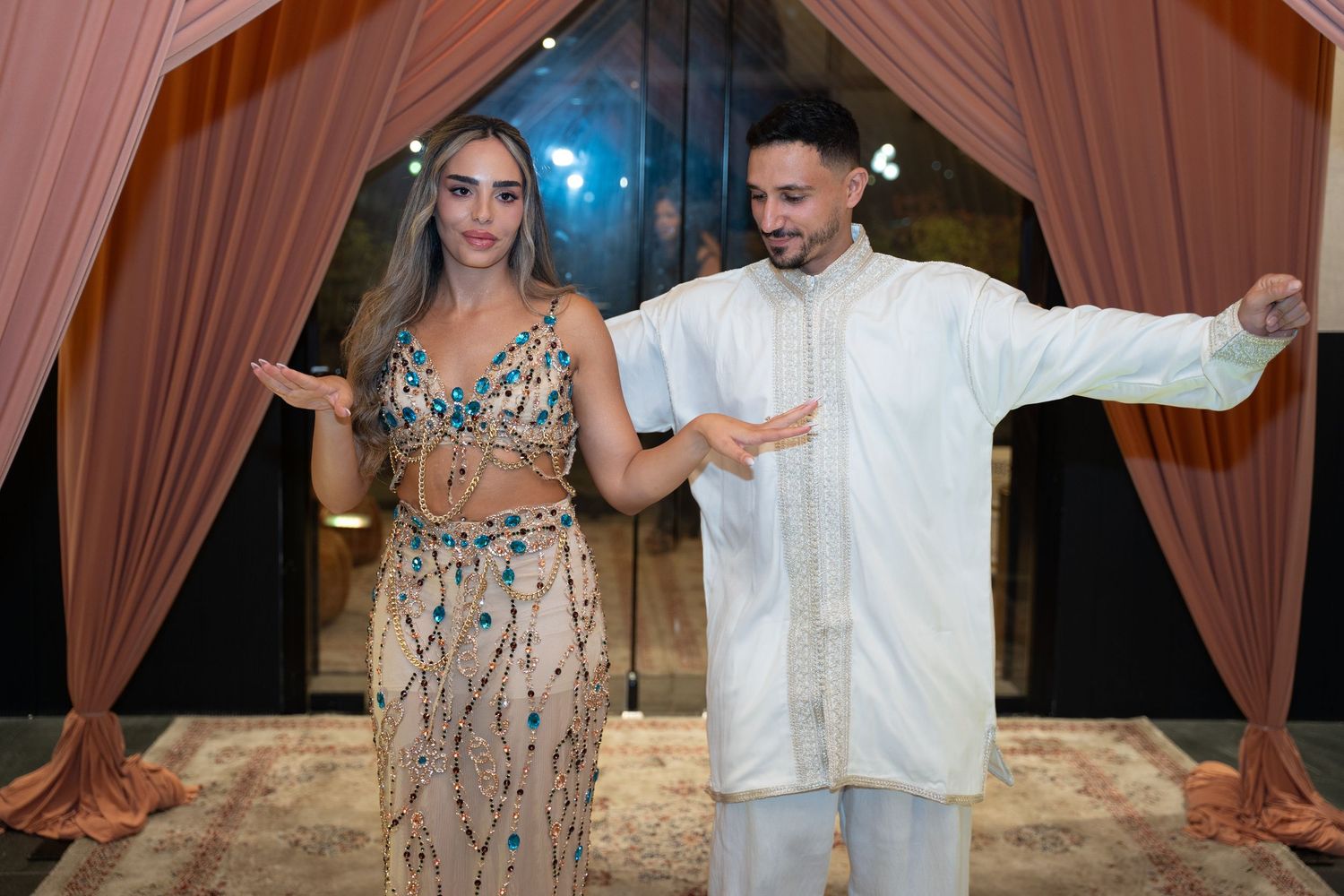 Couple in elegant attire posing under pink drapes at a formal event, woman in beaded gown, man in white kurta.