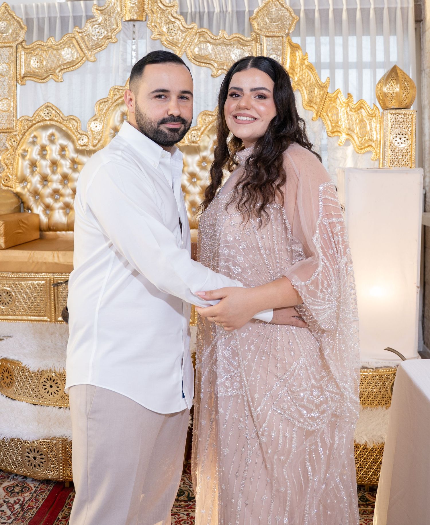 Couple in white and blush outfits posing against ornate gold backdrop.