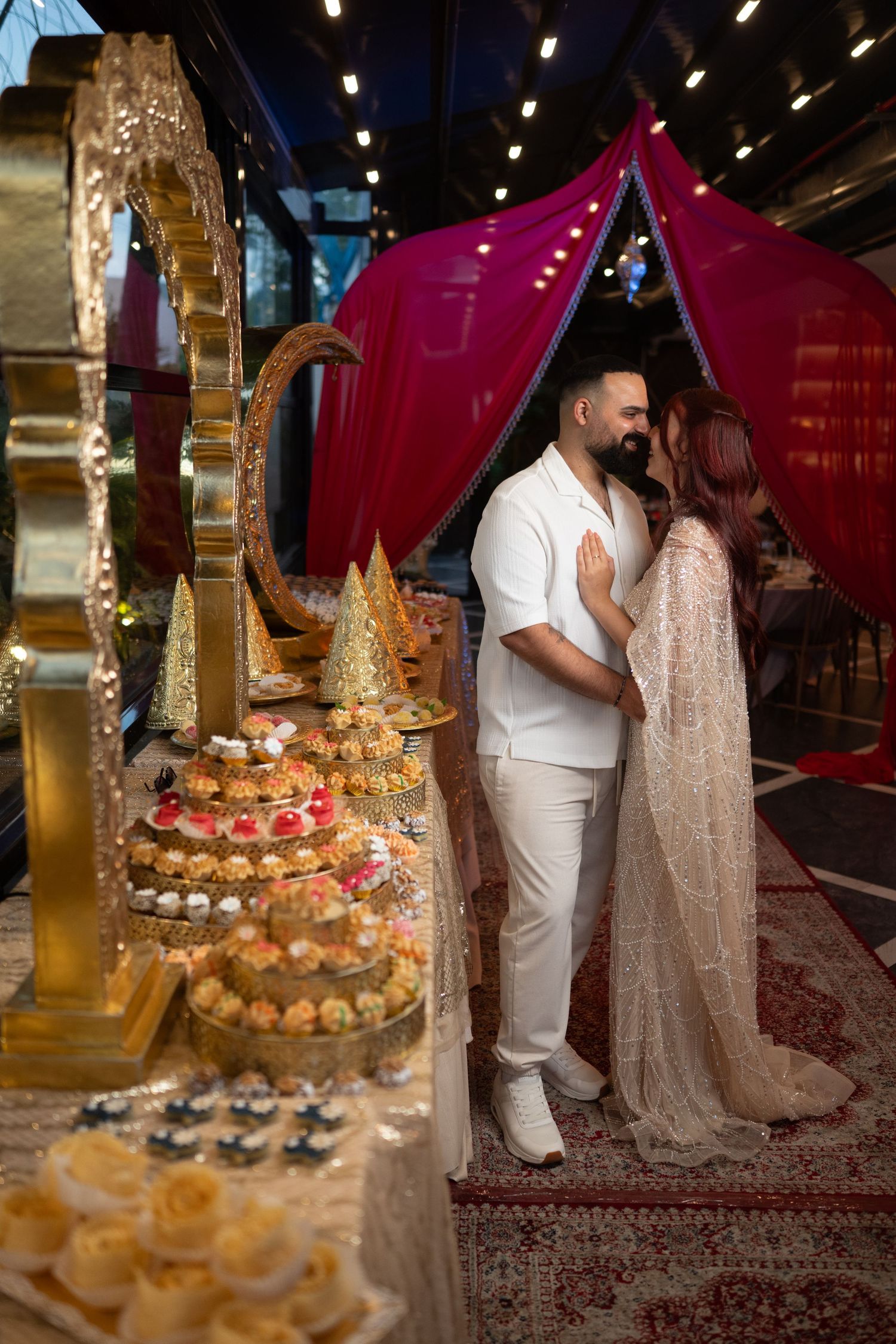 Couple in white attire sharing romantic moment near ornate wedding cake display with golden decor and red canopy backdrop.