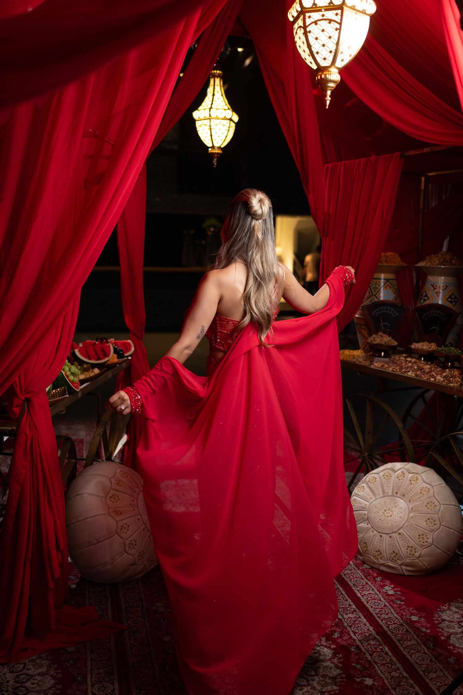 Woman in elegant red gown standing in ornate Moroccan-style room with red curtains and decorative lanterns.
