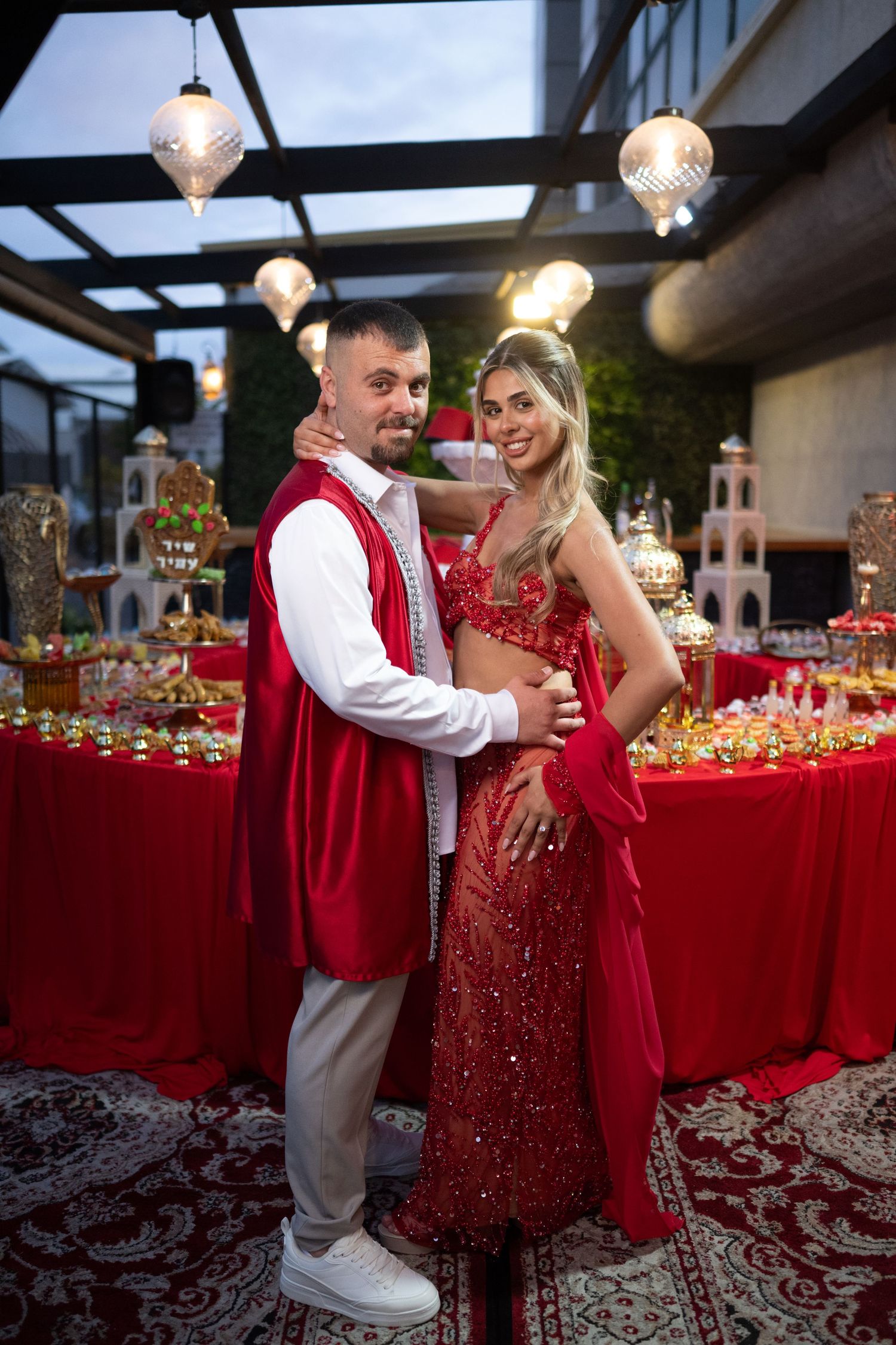 Couple in red traditional outfits posing at elegant event with decorative lights and candy table backdrop.