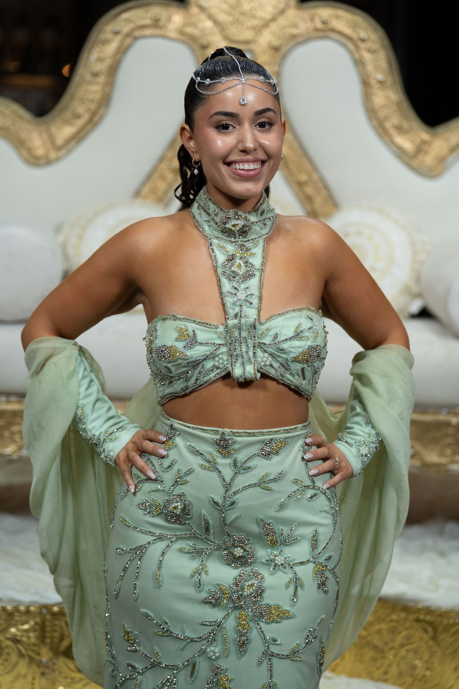 Woman in mint green embroidered lehenga with head jewelry smiles posing in front of ornate white and gold throne.