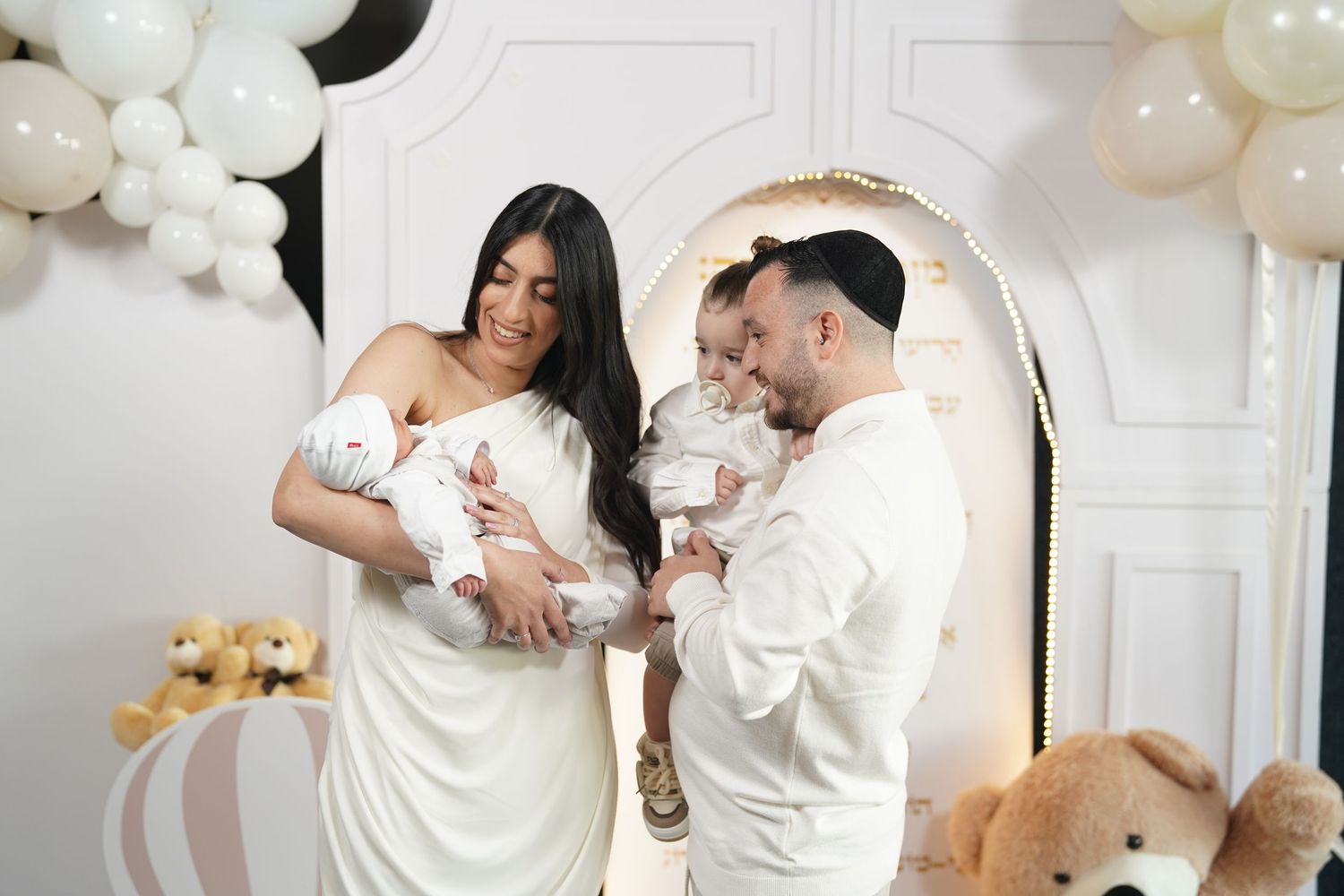 A newborn photoshoot setup with white balloons, decorative blocks, and a teddy bear against a white backdrop.