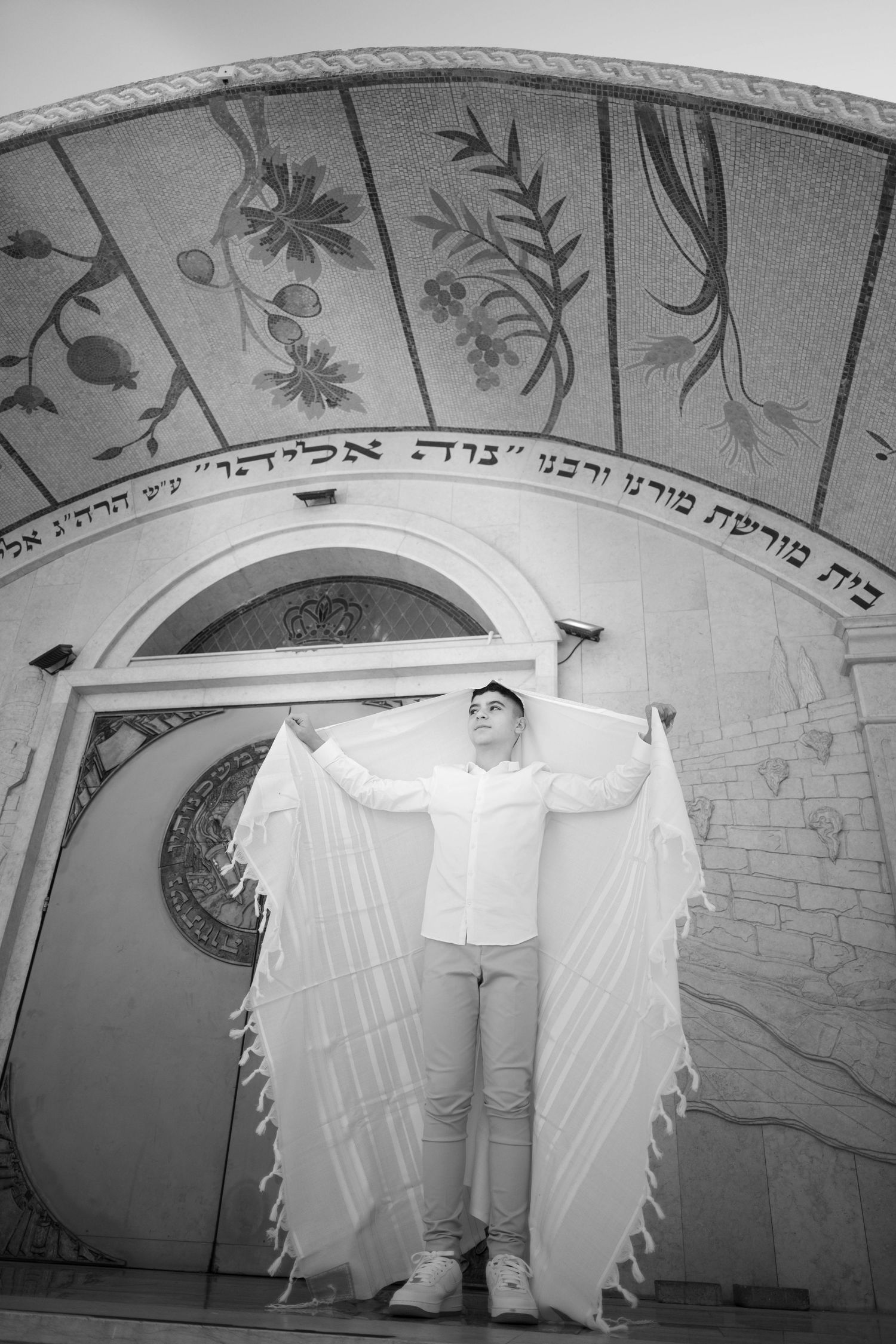 A person poses with white fabric wings against an ornately decorated archway with Hebrew text.