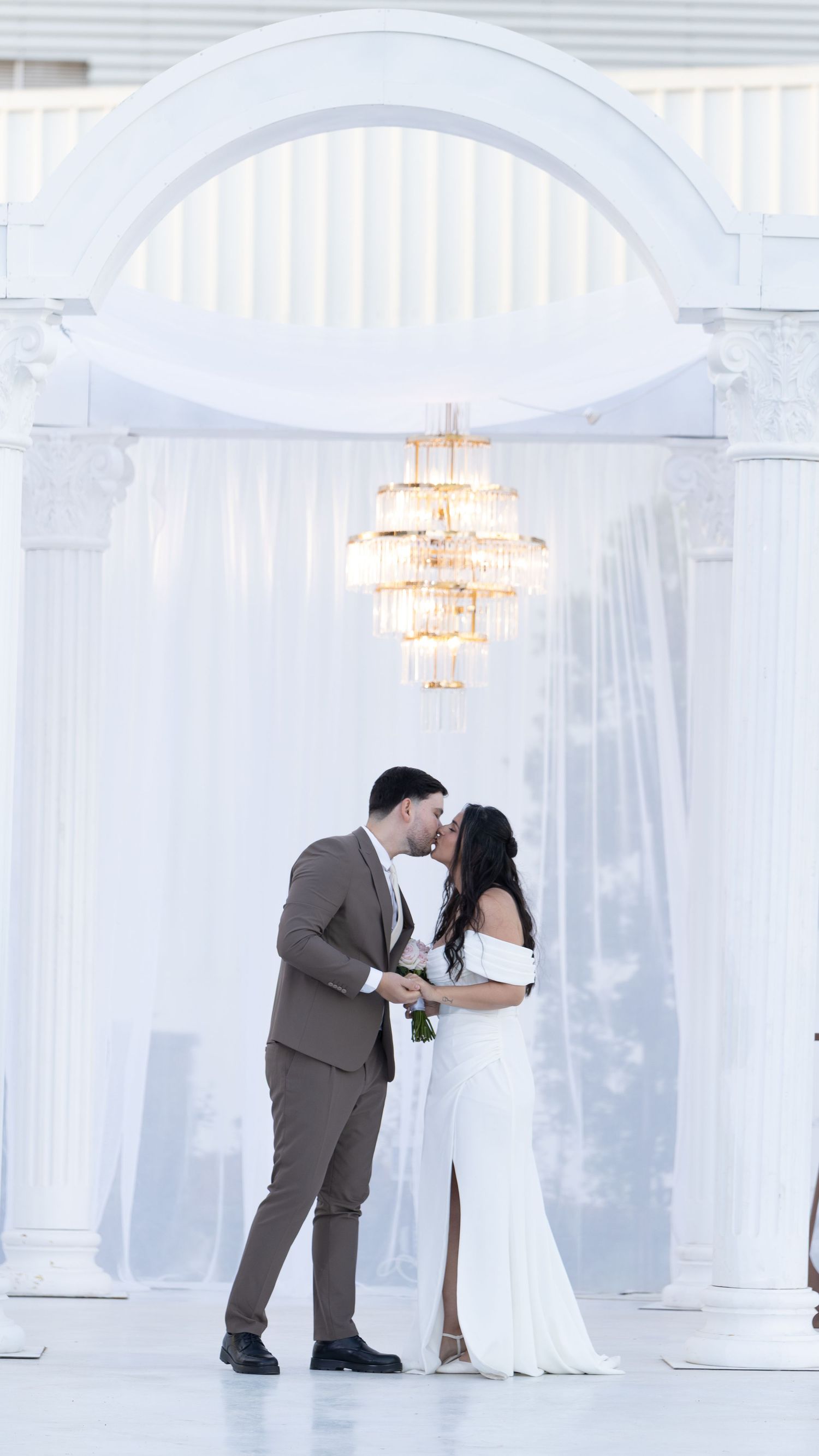 Couple sharing first kiss at elegant white wedding ceremony under chandelier and arch with draped fabric backdrop.
