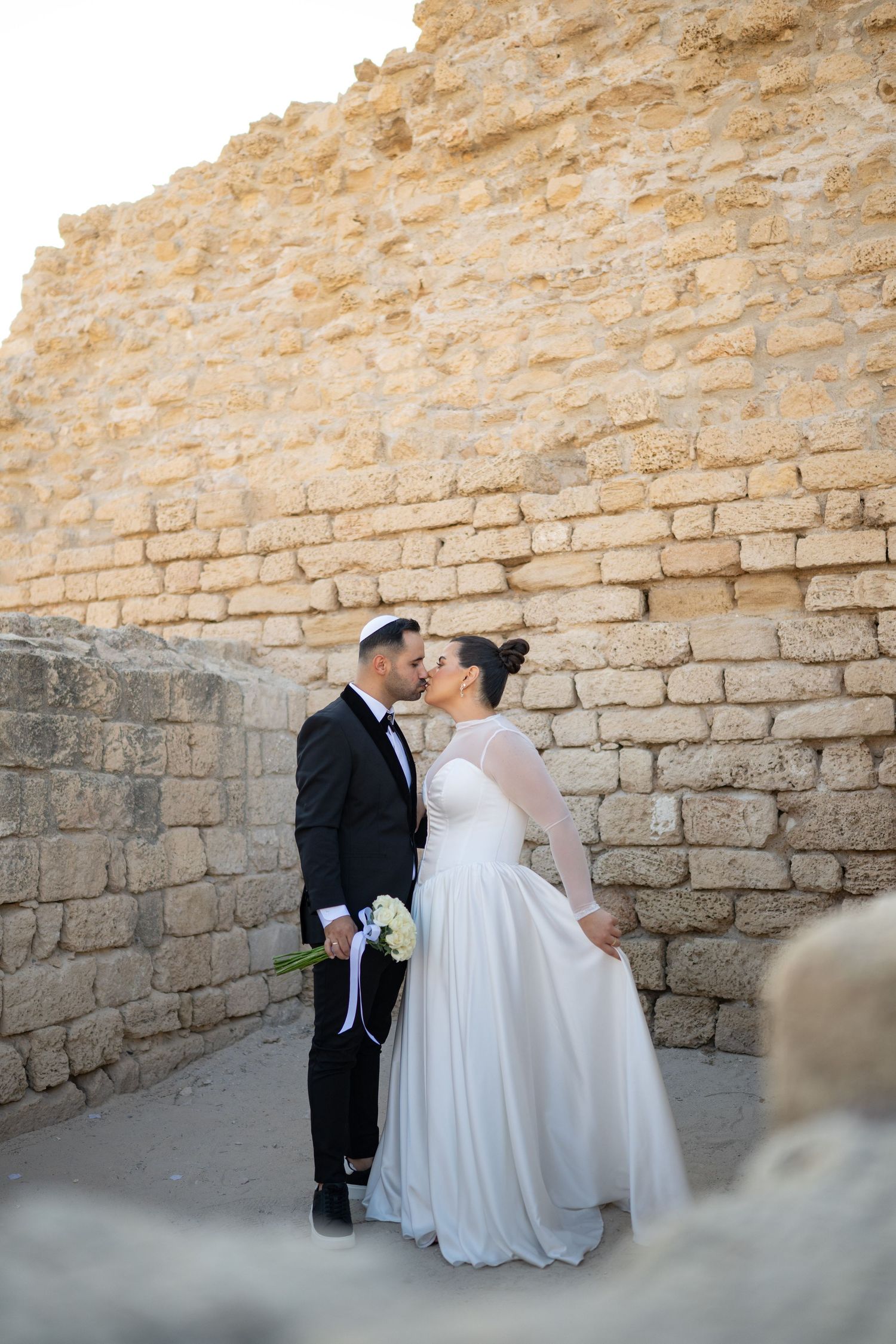 Wedding couple poses against rustic stone wall in natural lighting.