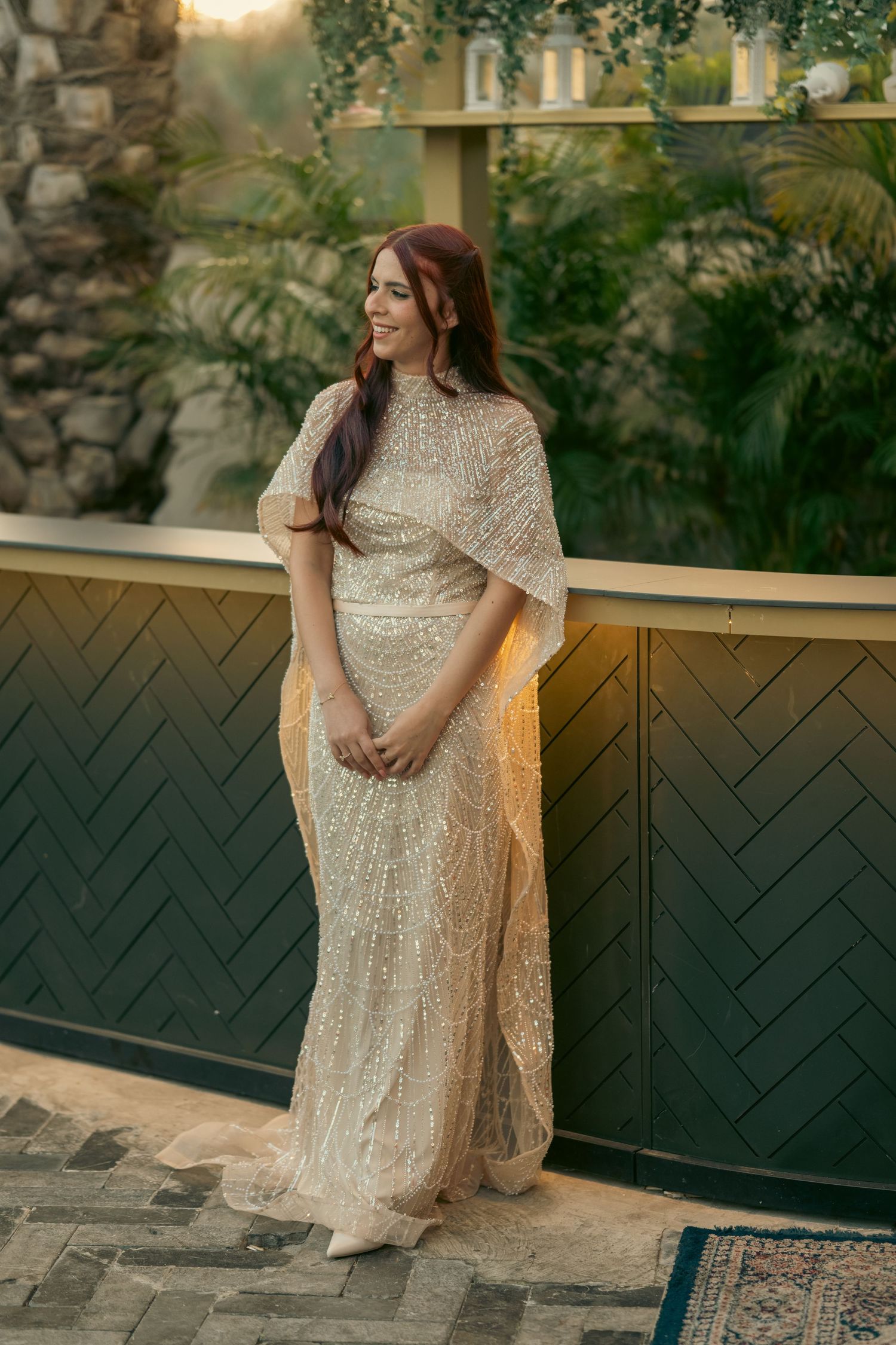 Woman in elegant sequined cream gown with cape sleeves poses outdoors at golden hour near lush greenery.