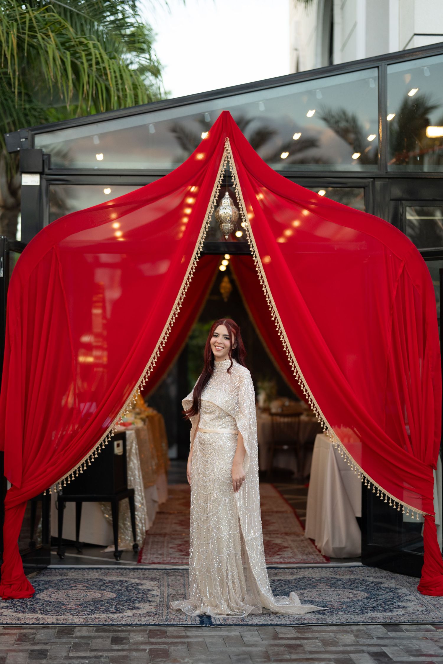 Bride in white lace gown stands under elegant red draped canopy with gold trim at a wedding venue.
