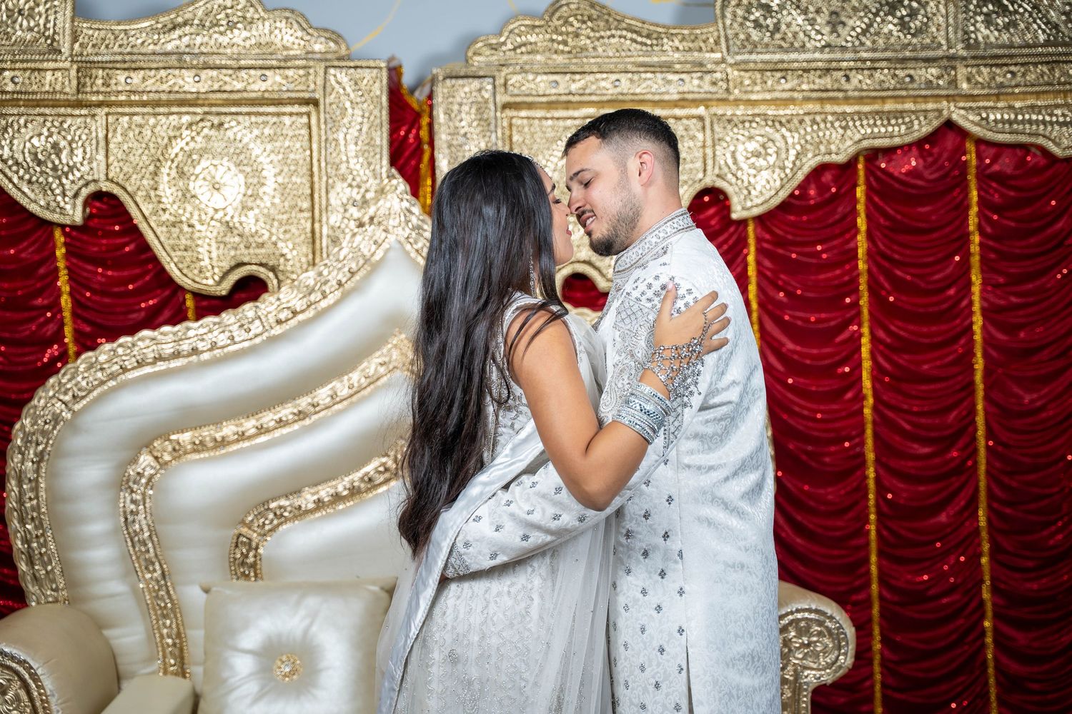 Elegant couple in white attire posing against ornate gold backdrop.