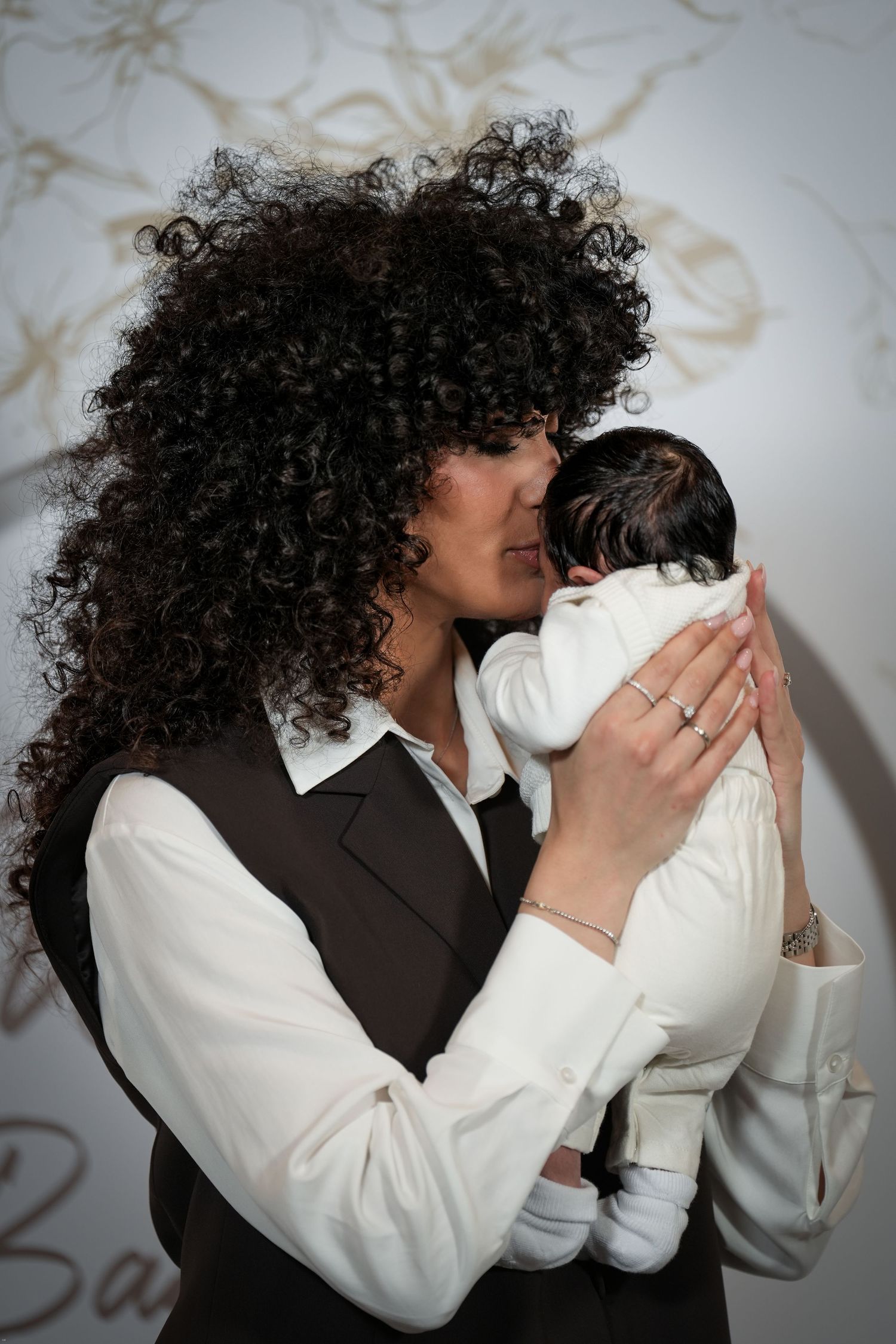 A woman with curly hair tenderly holds and kisses a newborn baby dressed in white.