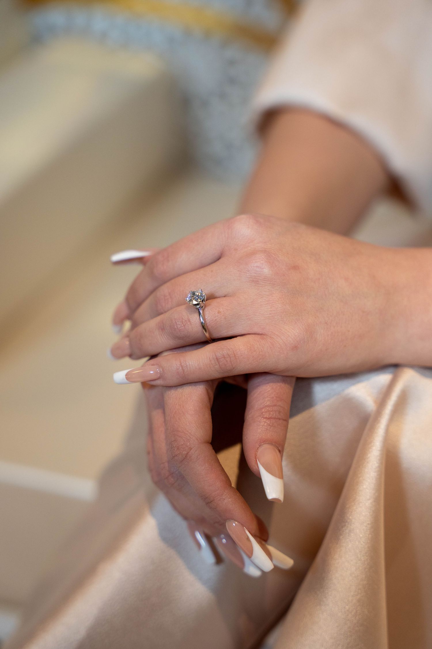 Close-up of hands with French manicured nails and a solitaire diamond engagement ring resting on champagne colored fabric.