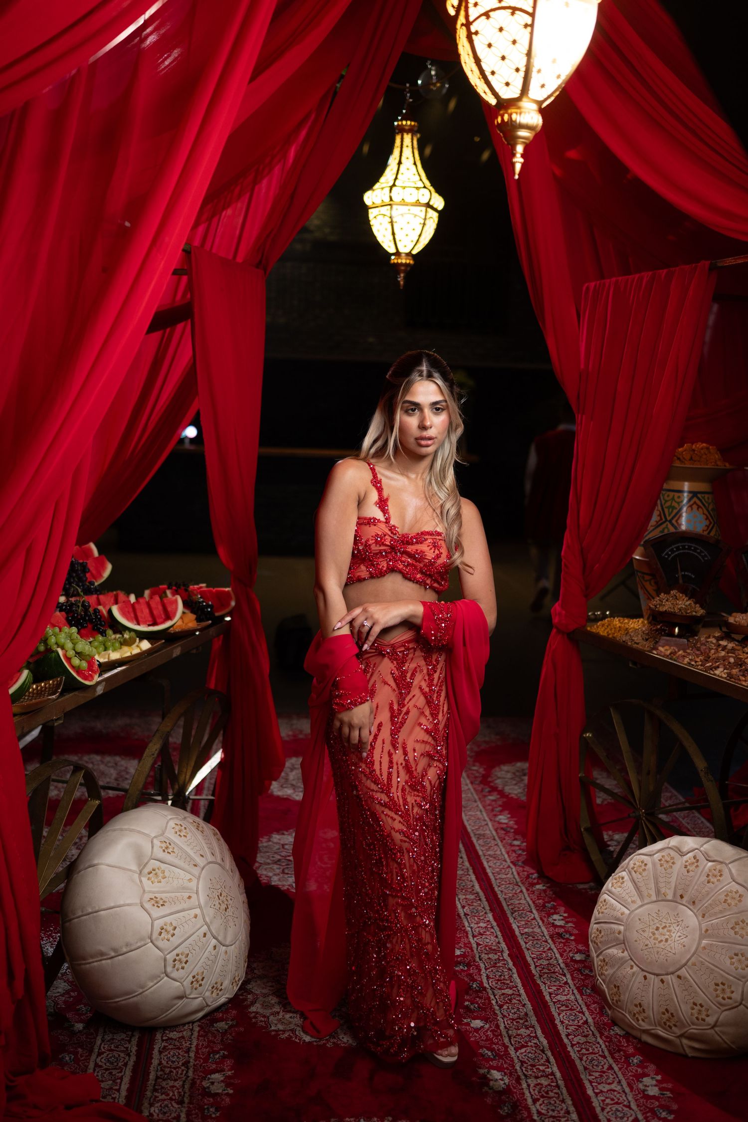 Woman in red sequined outfit poses under ornate lantern amid red drapes and Moroccan decor.